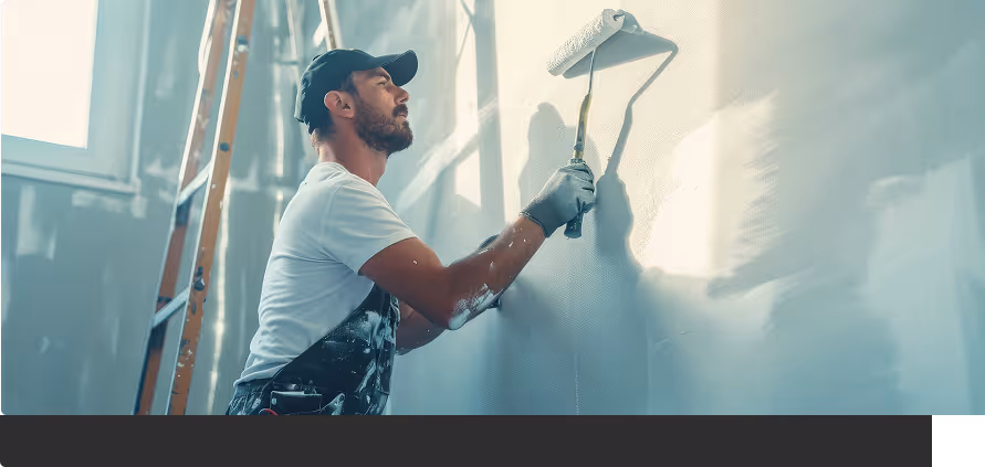 Mann in weißem T-Shirt und schwarzer Kappe streicht mit Farbwalze eine Wand in einem hellen Raum.