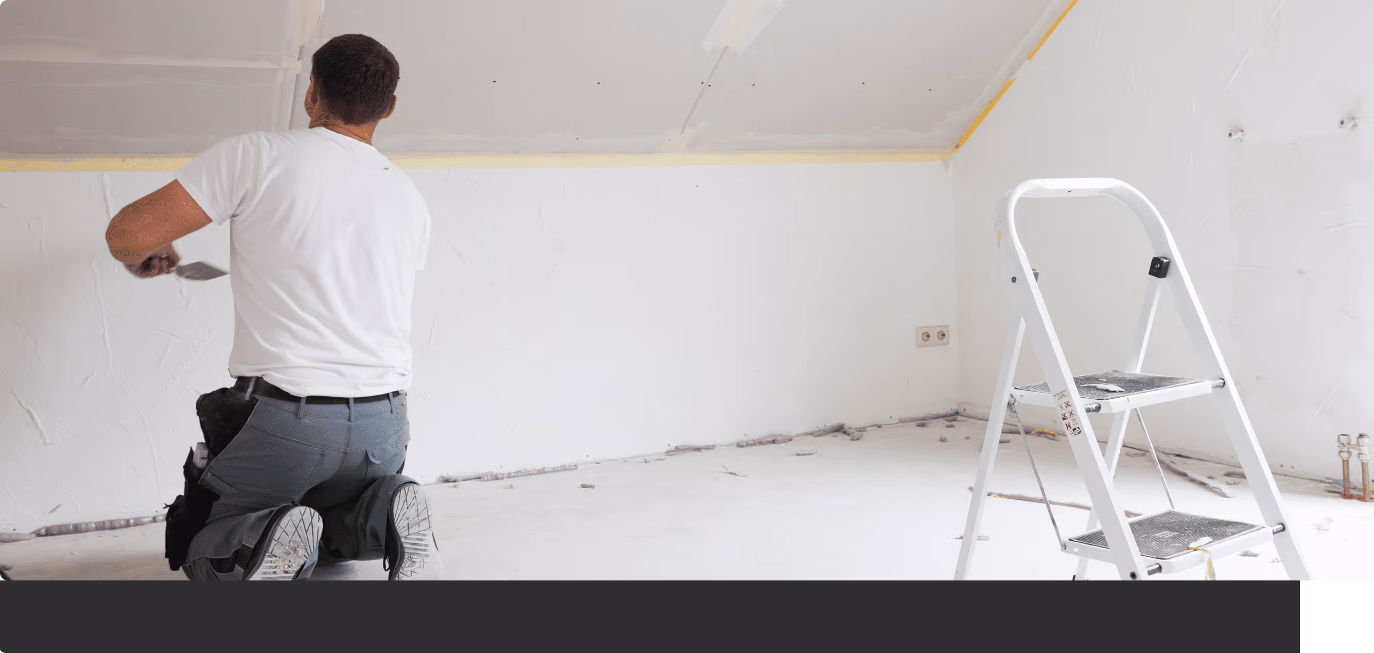 Mann kniet in einem leeren, weißen Raum und trägt Renovierungsarbeiten mit einem Spachtel aus, neben einer weißen Stehleiter.