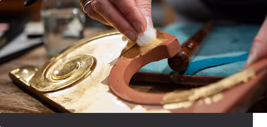Hand reinigt mit Watte einen vergoldeten kunstvoll verzierten Gegenstand auf einem Tisch.