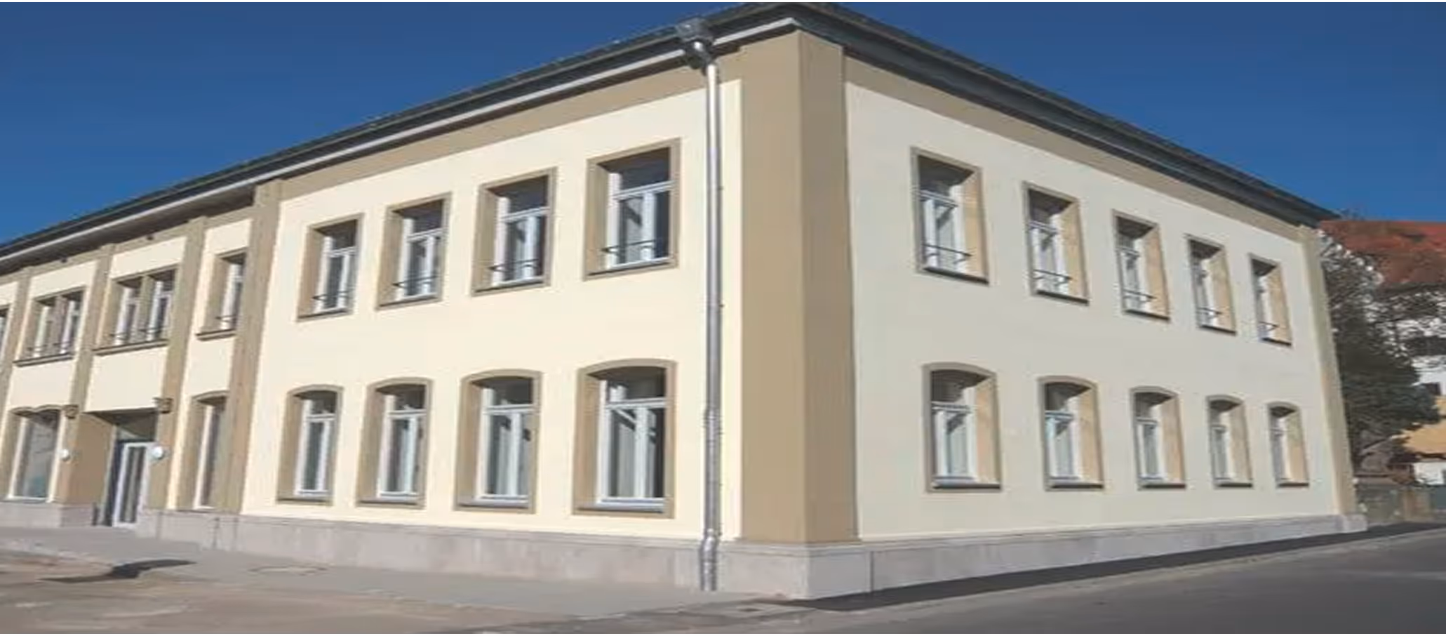 Zweistöckiges Gebäude mit gelblich-beiger Fassade und mehreren Fenstern an einem sonnigen Tag.