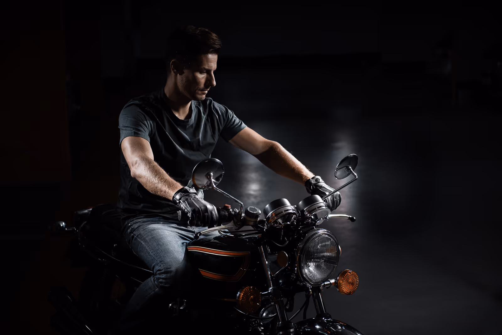 Man wearing black gloves and a black t-shirt sitting on a vintage motorcycle in a dimly lit setting.