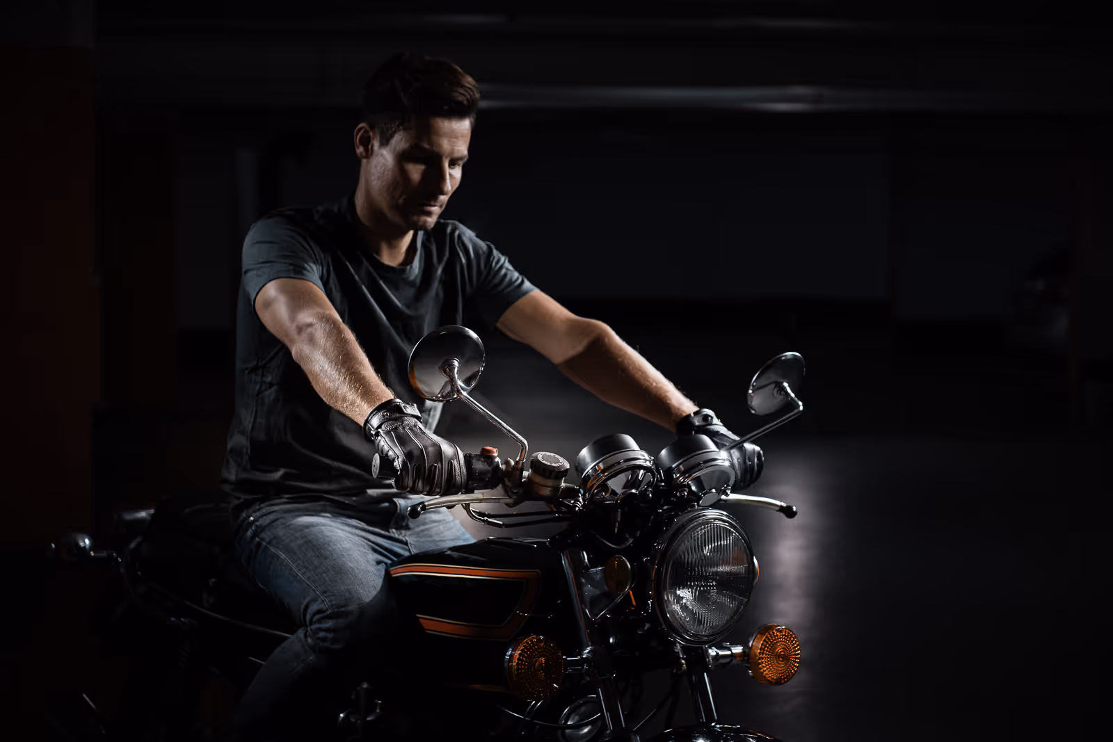 Man wearing black gloves and a dark t-shirt sitting on a classic motorcycle in a dimly lit setting.