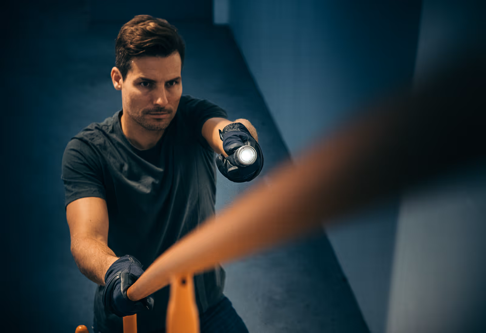 Man in black gloves holding a flashlight and a wooden baseball bat in a dimly lit corridor.