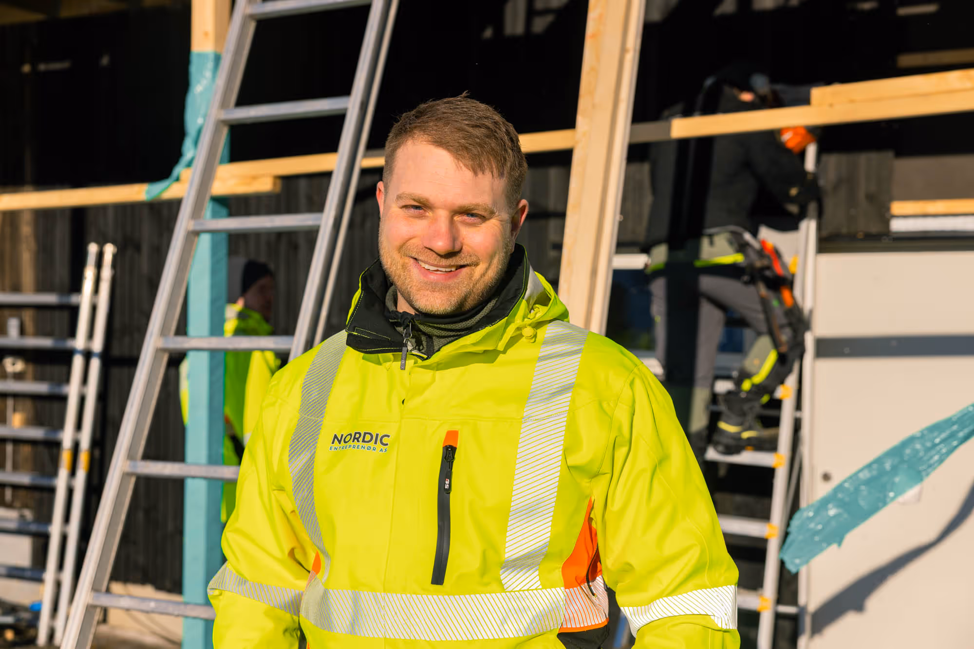 Smilende mann i gul høy synlighet jakke med reflekser på en byggeplass med stiger og arbeidsfolk.