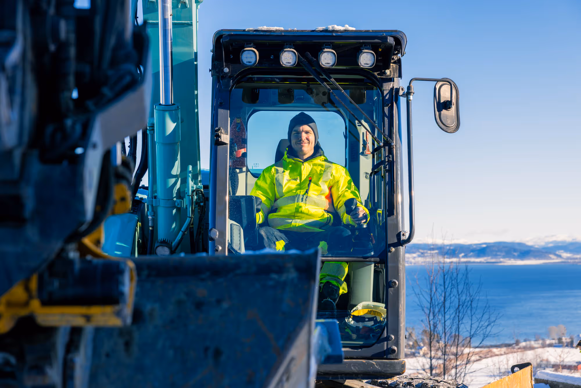 Mann i gul arbeidsjakke og lue sitter i førerhuset på en gravemaskin med snødekt landskap og sjø i bakgrunnen.