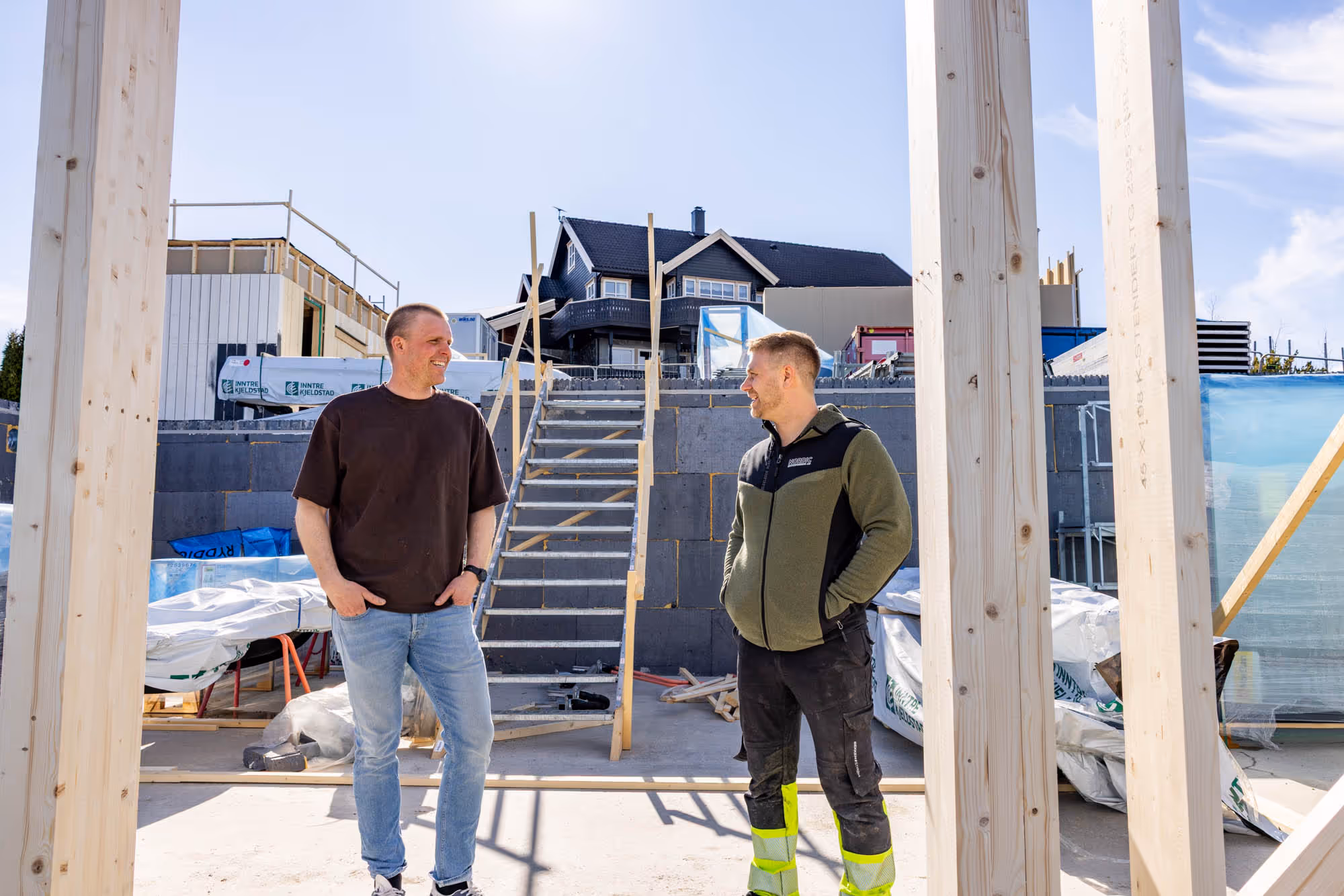 To menn i samtale på en byggeplass med trapp og hus under oppføring i bakgrunnen.