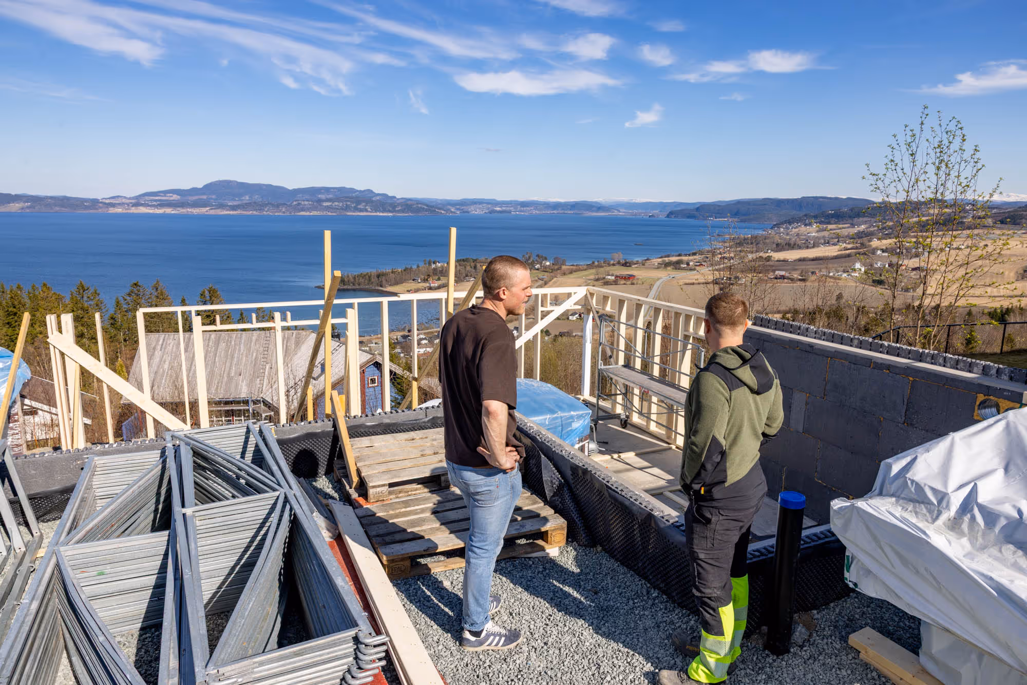 To mennester på en byggeplass med utsikt over innsjø, fjell og landsby i bakgrunnen.