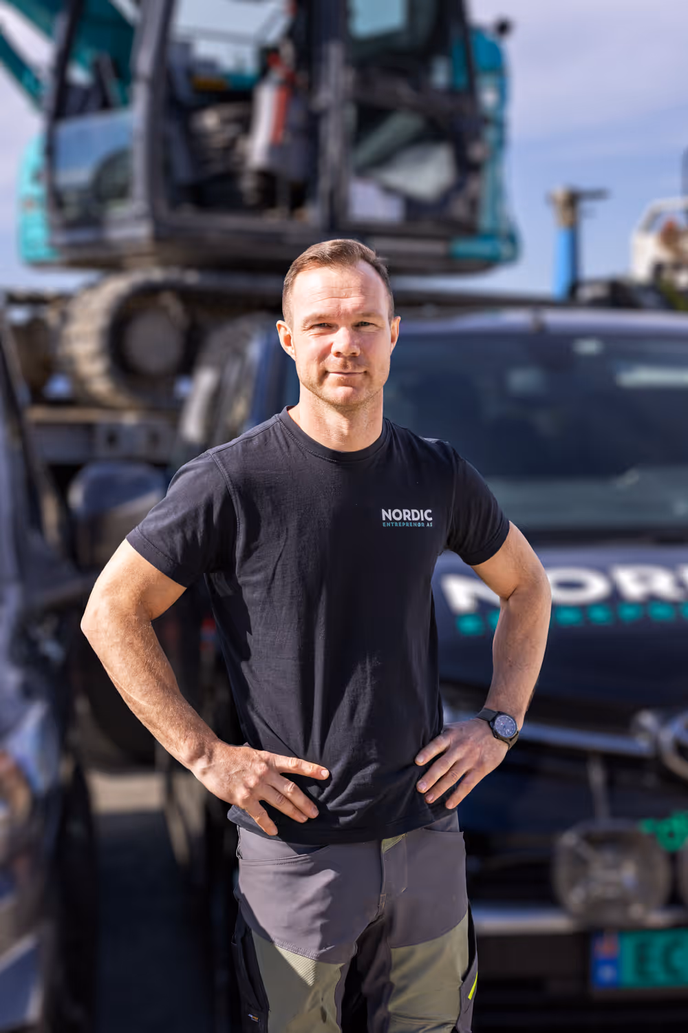 Mann i svart t-skjorte med Nordic Entrepreneur AS-logo står foran anleggsmaskin og varebil.