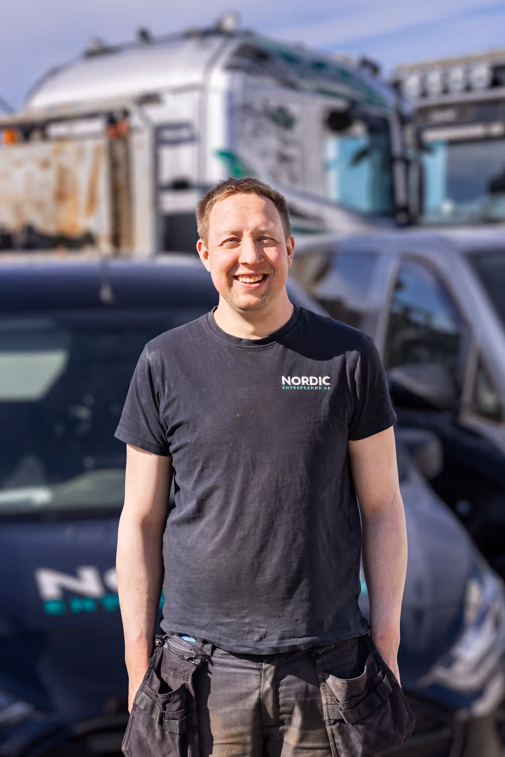 Smilende mann i svart Nordic Entrepreneur t-skjorte foran lastebil og bil i dagslys.