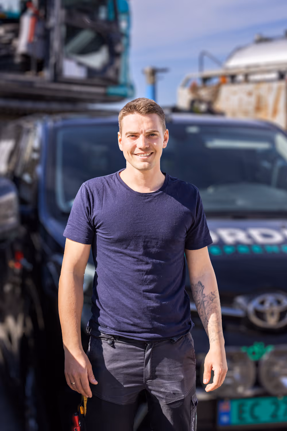 Ung mann med tatovering og blå t-skjorte smiler foran en svart Toyota varebil under sollys.