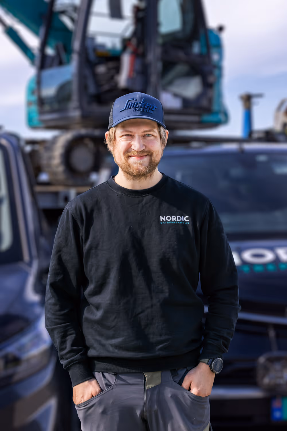 Smilende mann med sort skjorte og caps står foran arbeidskjøretøy i en byggeplass.