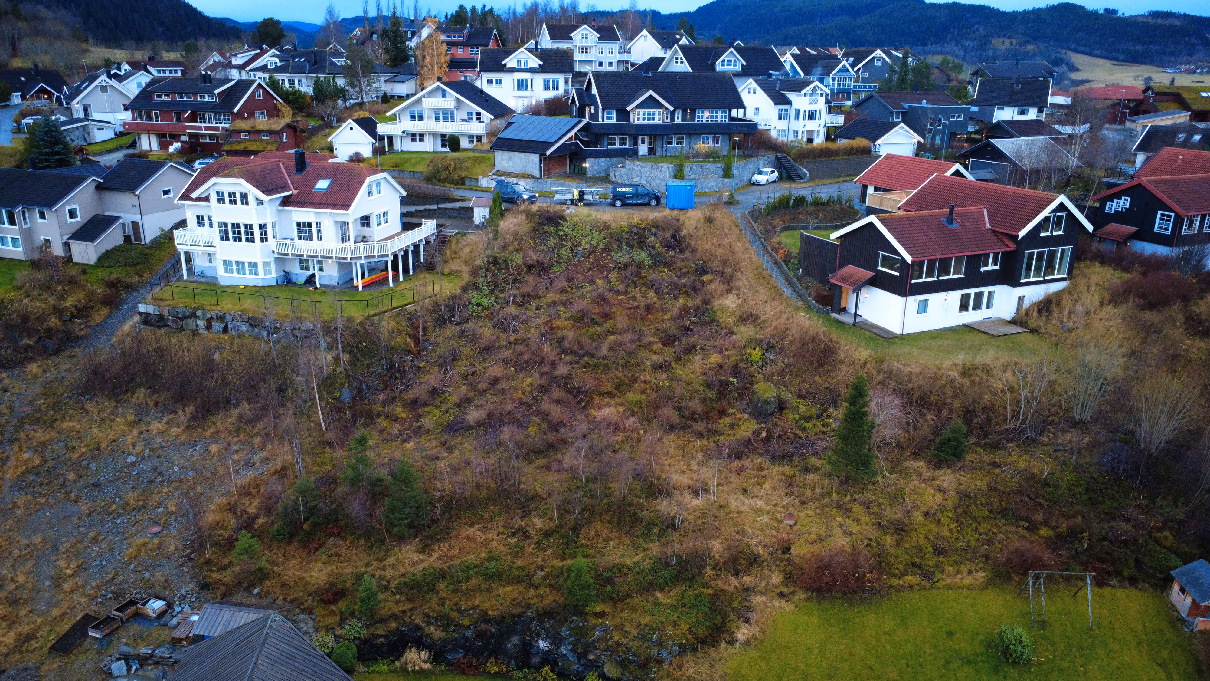 Boligområde med hvite og røde hus ved skogkledde åser under blå himmel.