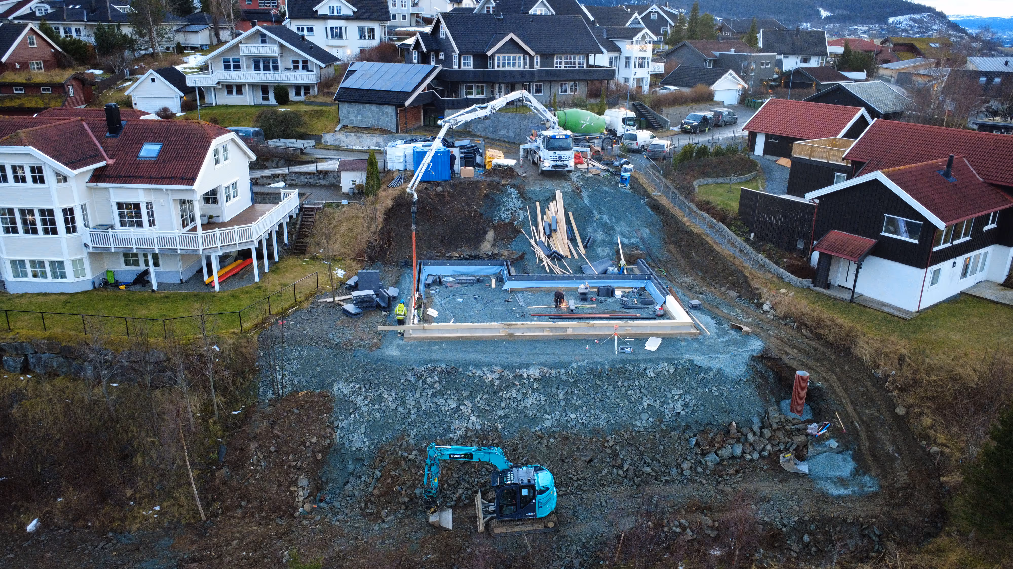 Byggeplass mellom hus med gravemaskin, betongbil og arbeidere under huskonstruksjon.
