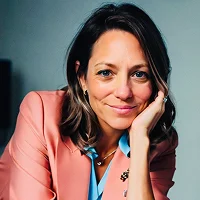 Woman with shoulder-length brown hair wearing a pink blazer and blue shirt, smiling and resting her face on her hand.