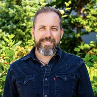 Smiling man with a beard wearing a dark denim shirt standing outdoors with green foliage in the background.