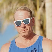 Man wearing white sunglasses and a blue tank top standing outdoors with blurred trees and sky in the background.