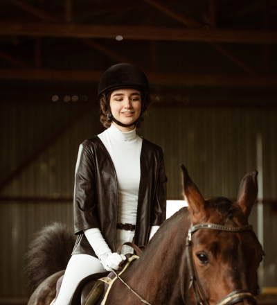 Young woman in equestrian gear riding a brown horse indoors with a calm expression.