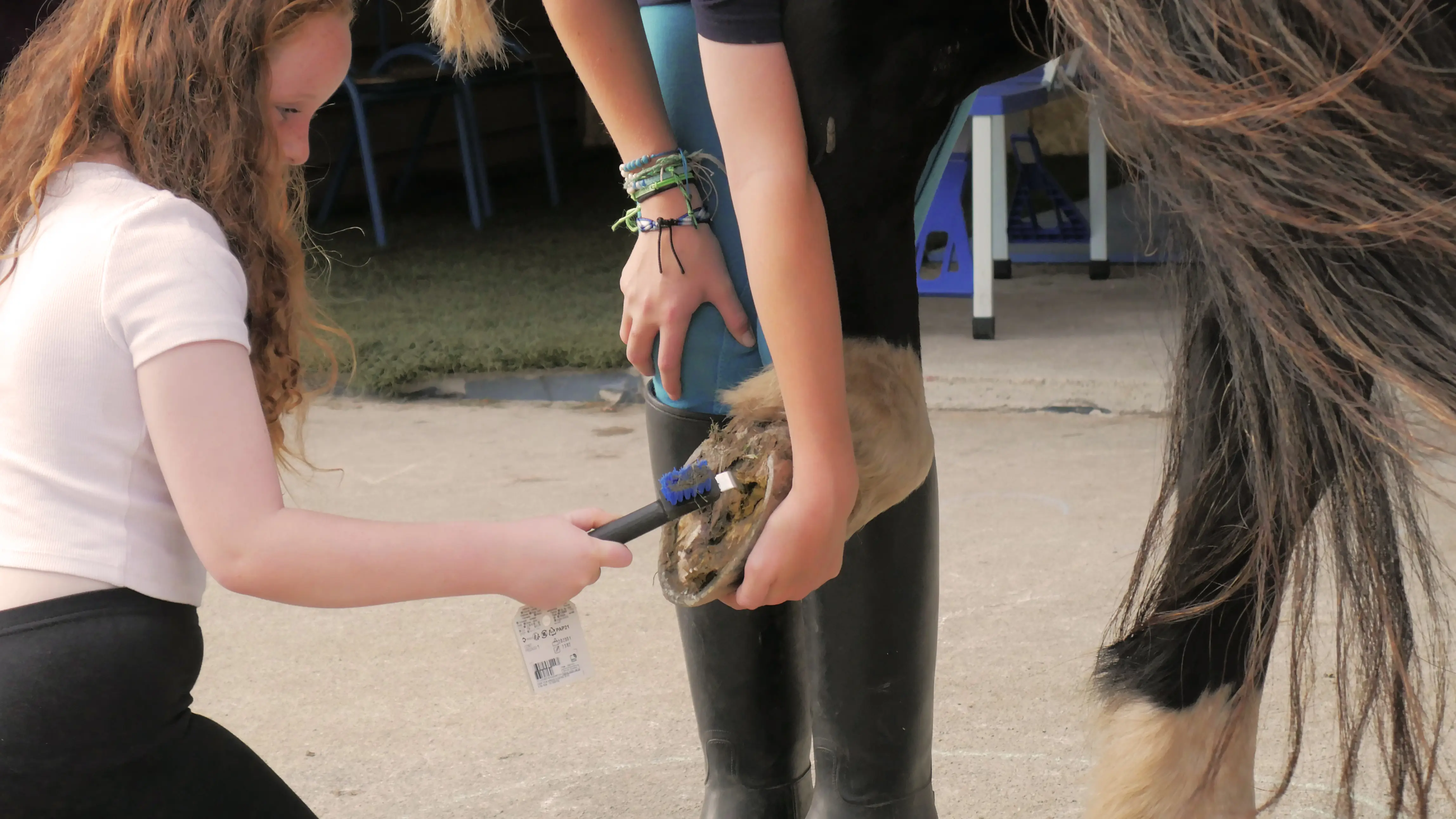 A child brushing the muddy hoof of a horse being held by another person wearing riding boots.