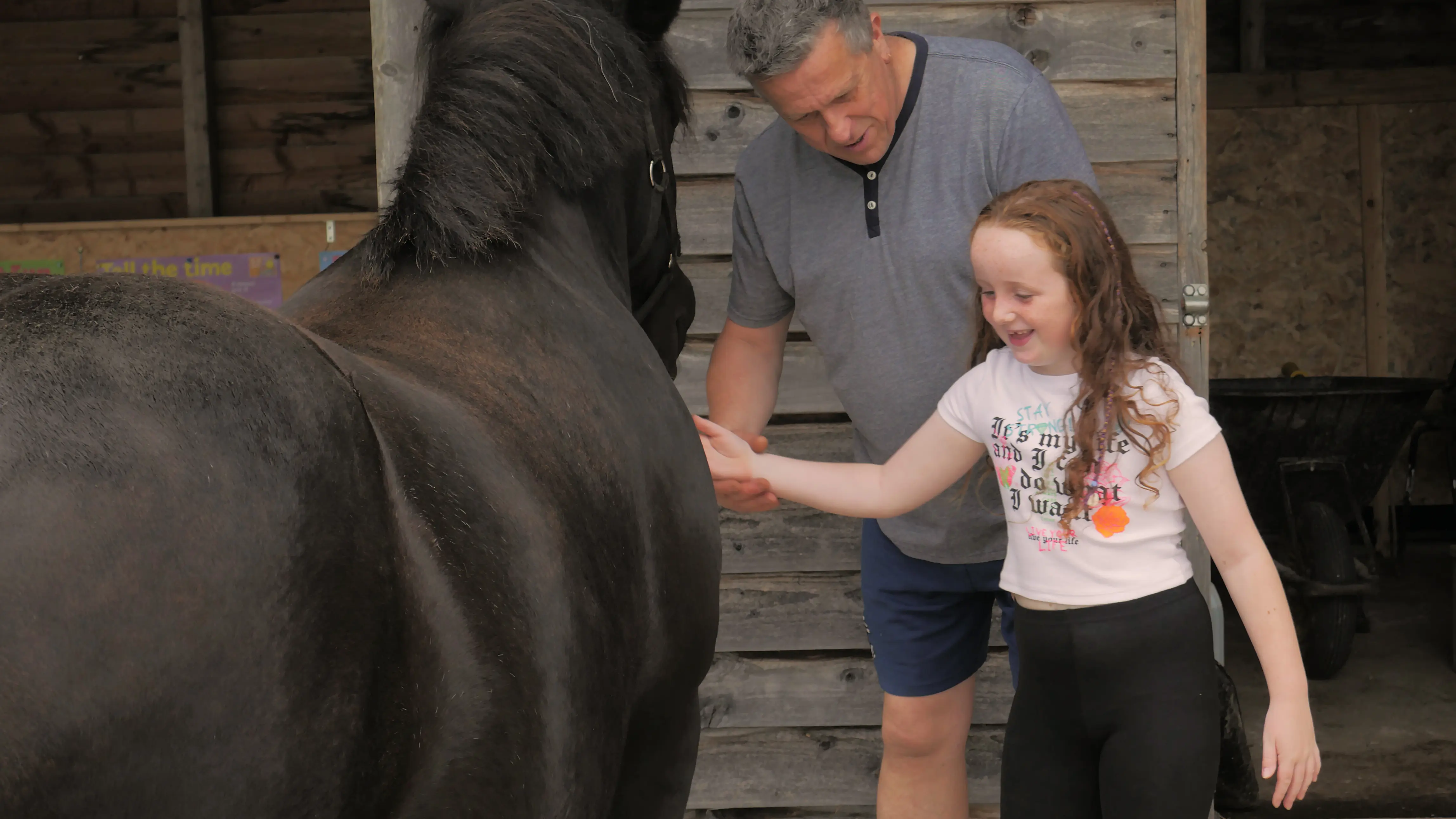 Man and smiling girl petting a black horse in front of a wooden stable.