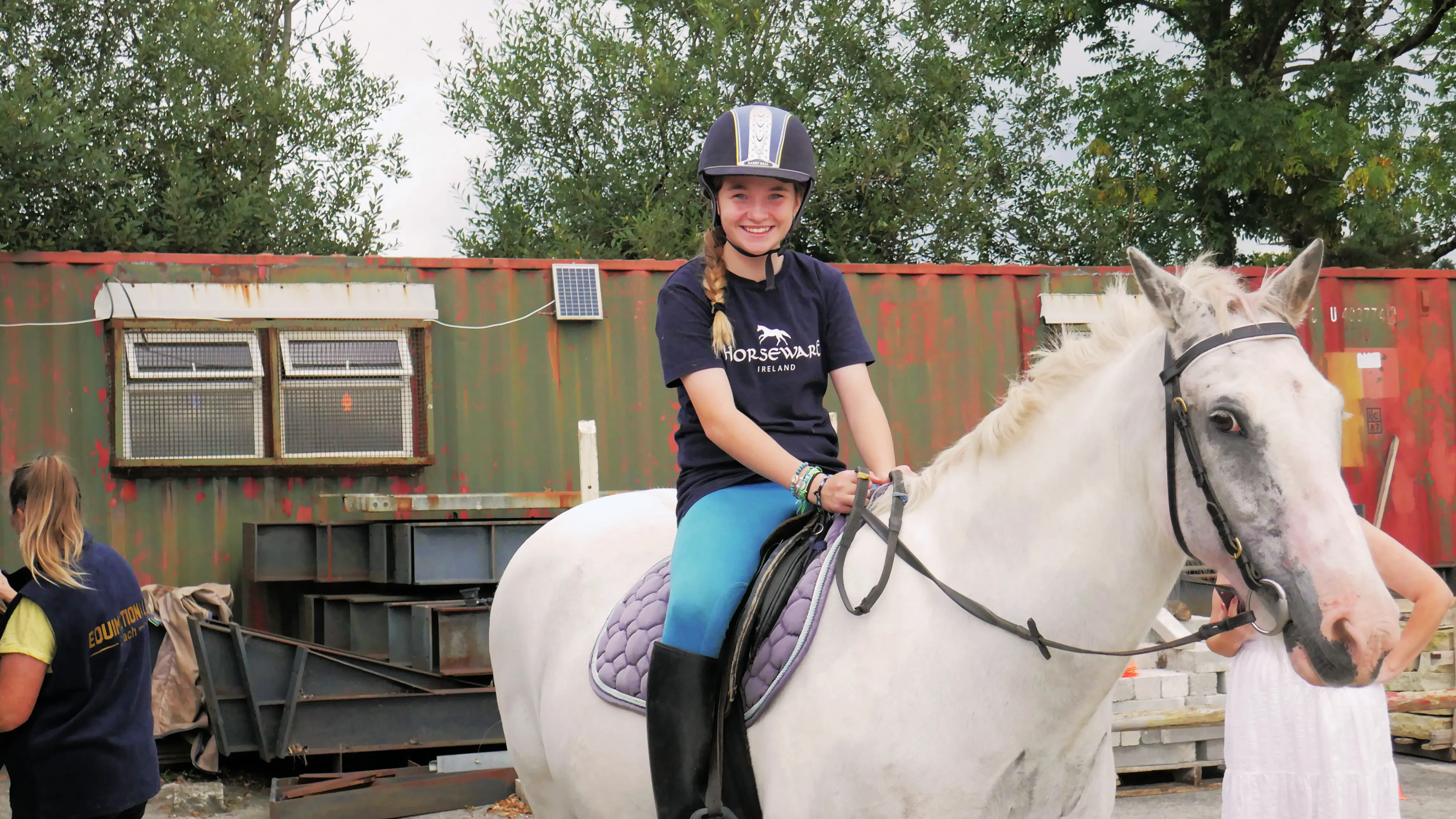 Smiling girl wearing a helmet riding a white horse with a purple saddle pad in an outdoor area with trees and a metal container in the background.