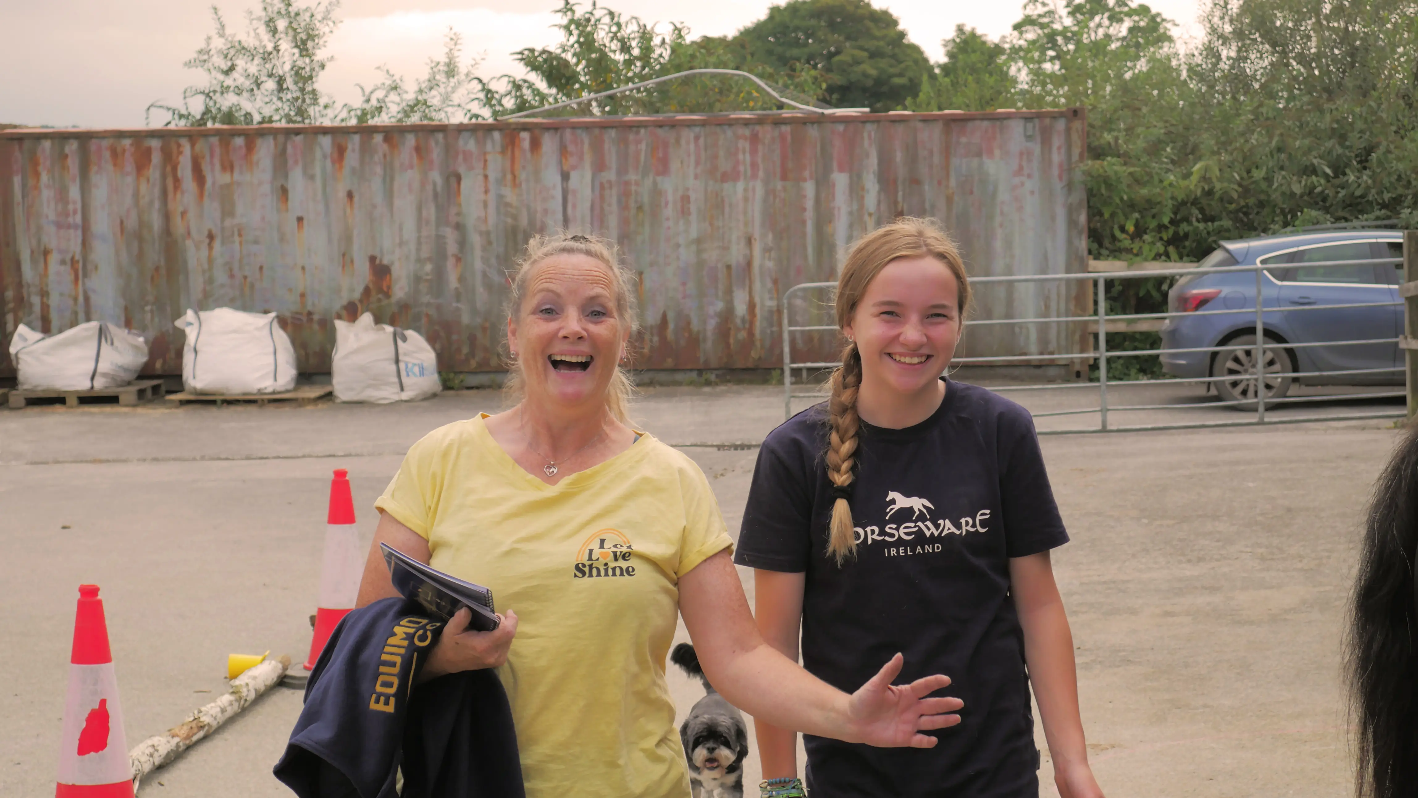 Two smiling women standing outdoors in front of a rusty shipping container, one wearing a yellow Love Shine shirt and the other a navy Horseware Ireland shirt.