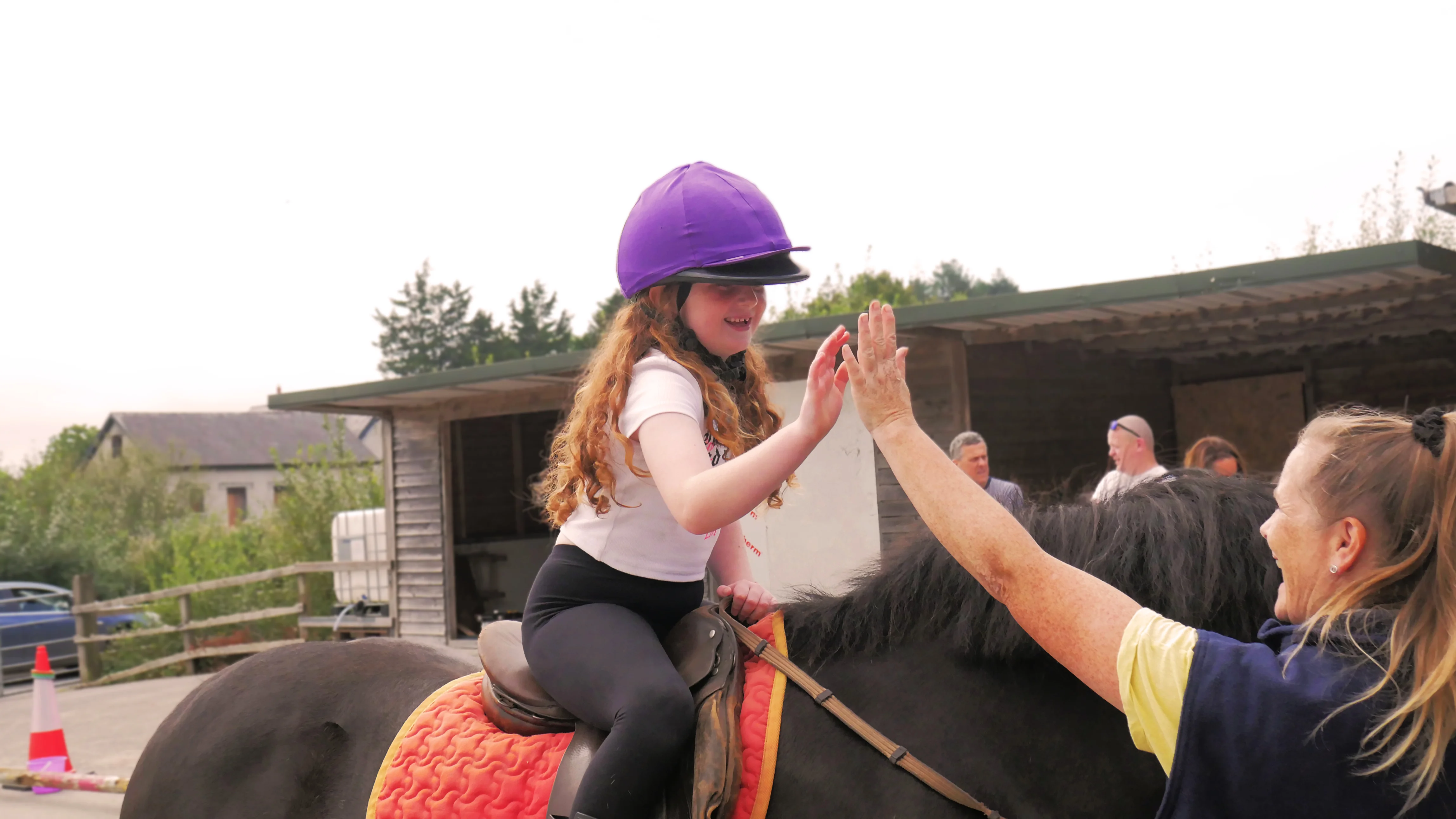 Smiling young girl wearing a purple helmet giving a high-five to a woman while sitting on a horse.
