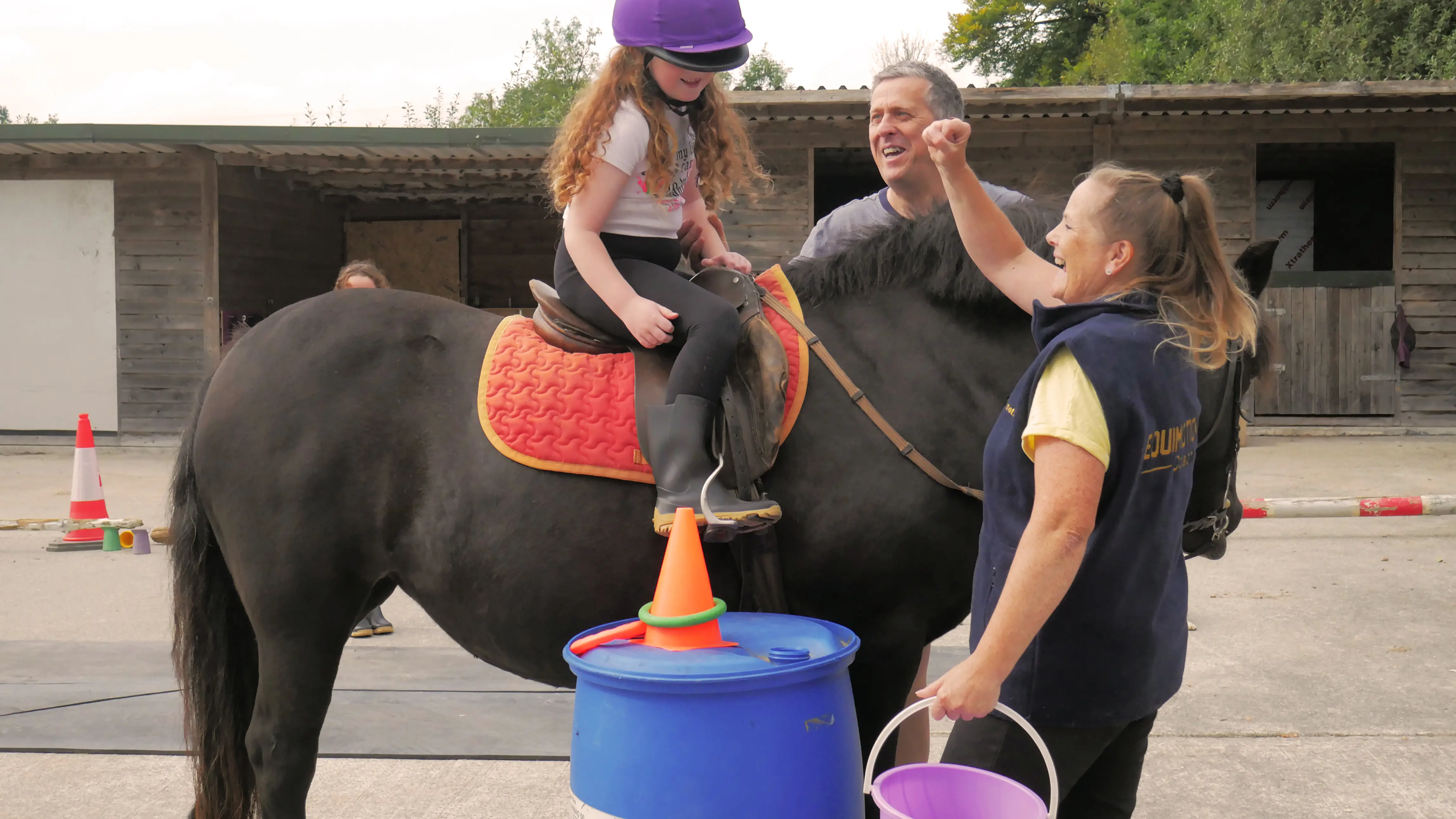 A young girl wearing a purple helmet sits on a black horse with an orange saddle pad while two adults encourage her during a riding lesson outdoors.