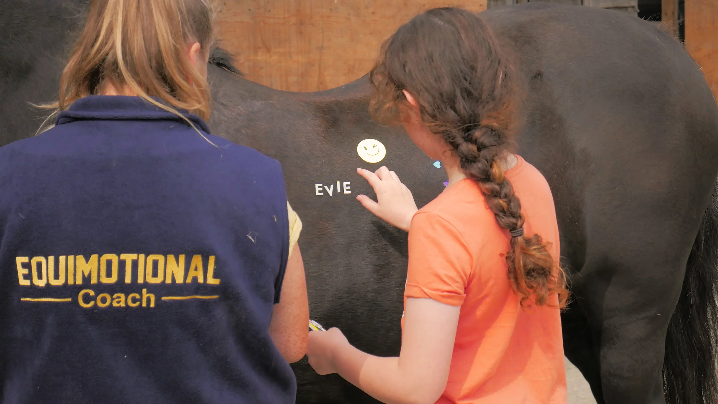 Two girls, one wearing an Equimotional Coach vest, placing letter stickers and a winking smiley face sticker on a black horse's side.