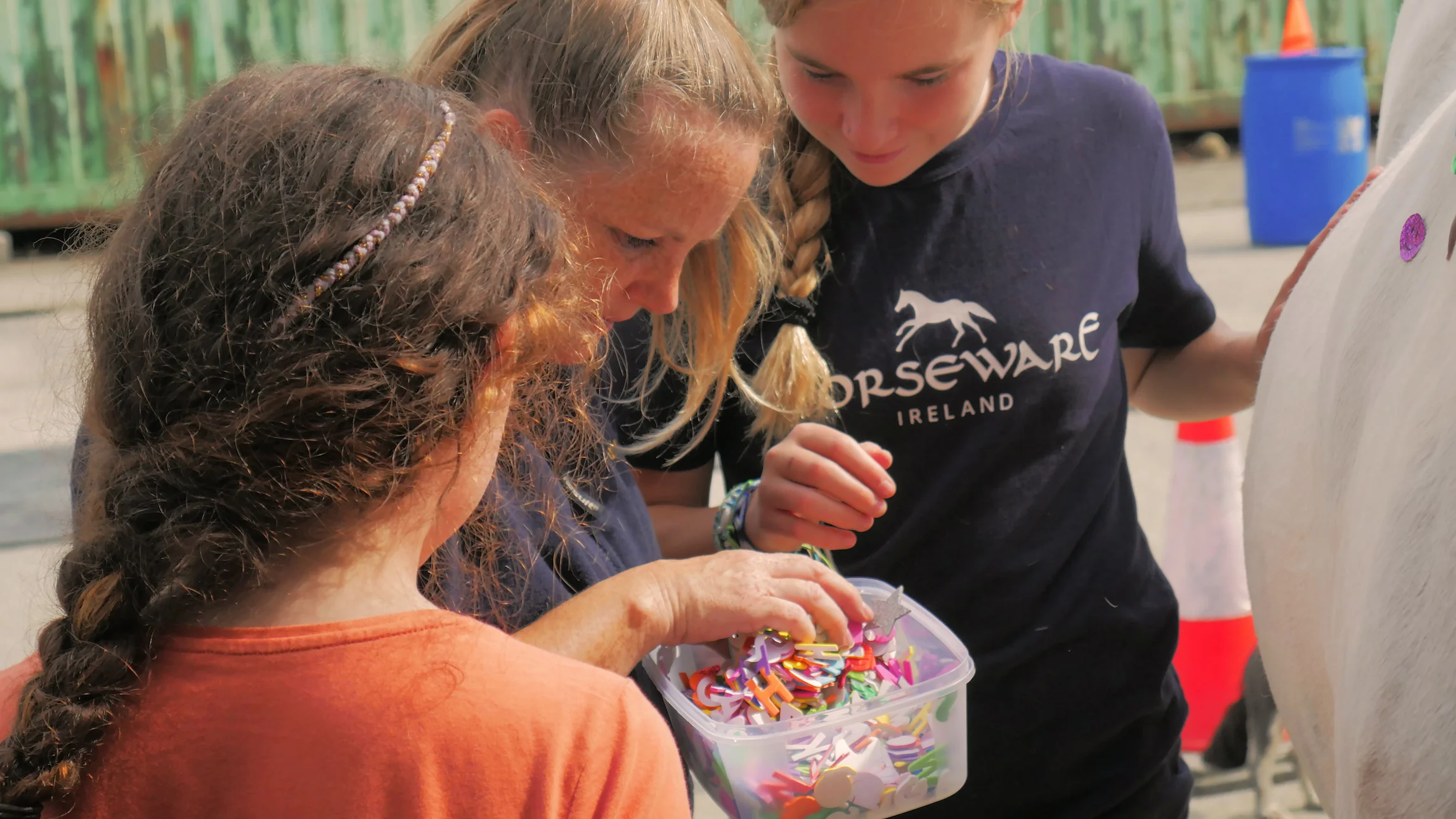 Three people looking into a container filled with colorful foam letters and shapes, standing next to a white horse.