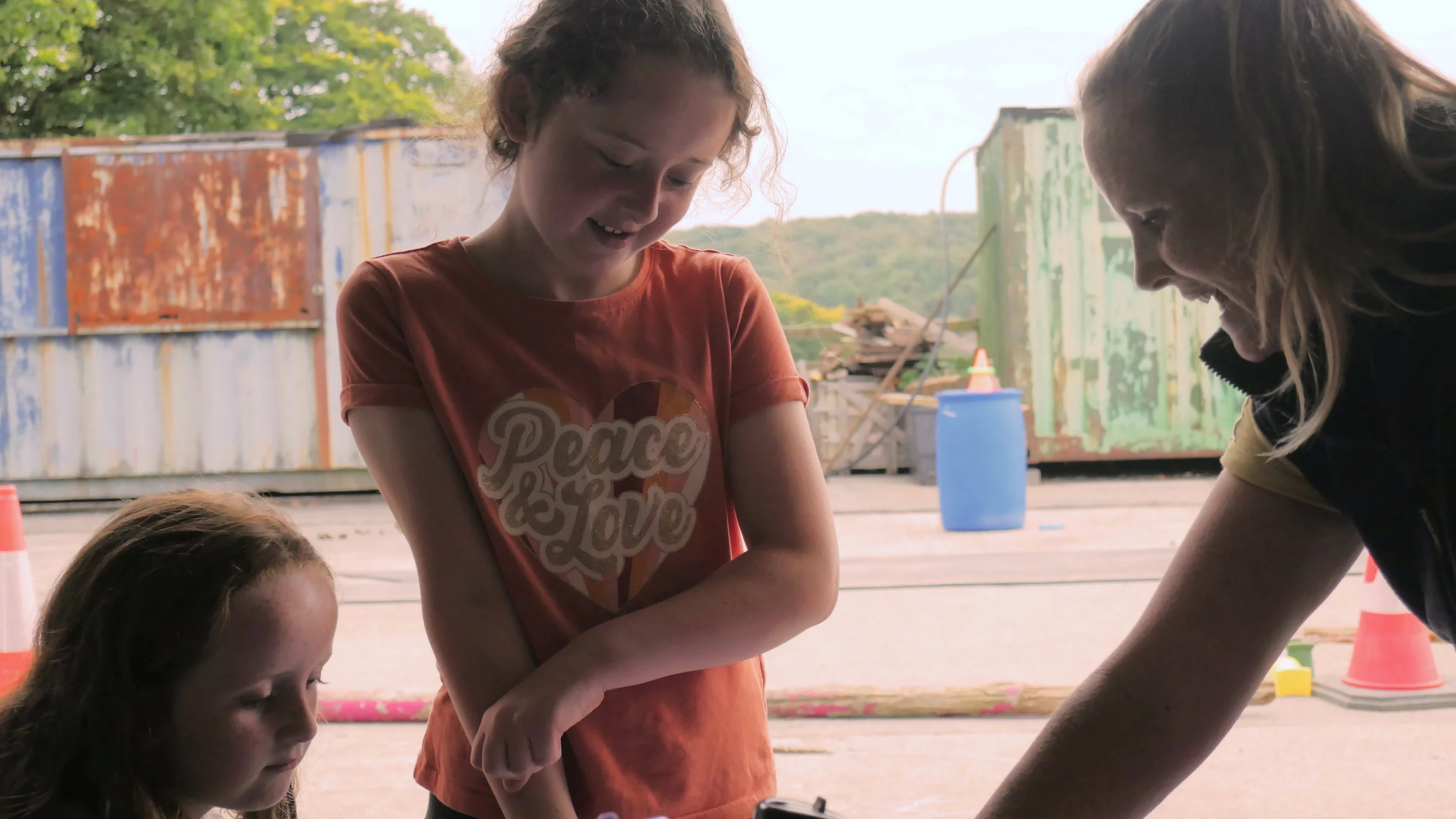 Two young girls interacting with a smiling woman outdoors near a rusted shipping container and traffic cones.