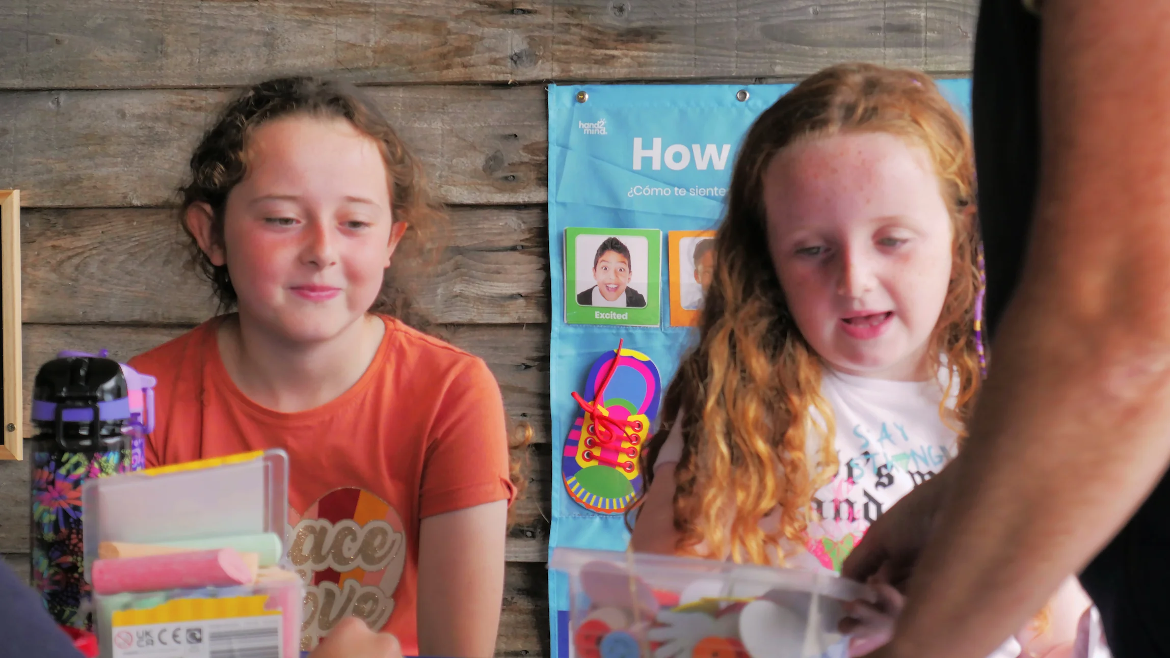 Two young girls sitting at a table with colorful chalk and craft supplies, looking at an adult's arm reaching toward them.