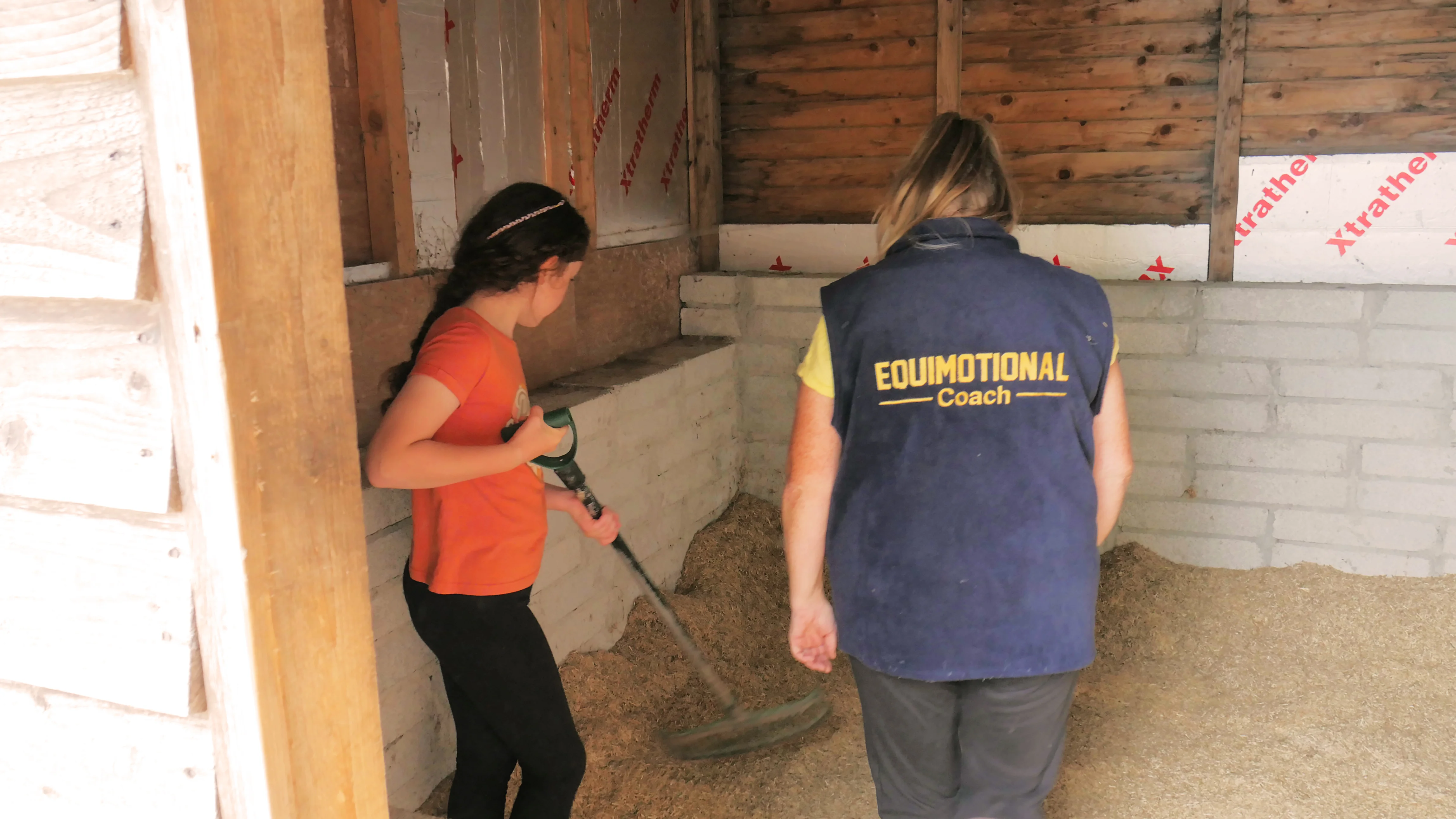 A girl with a rake and a person wearing an Equimotional Coach vest cleaning a stable with wood shavings on the ground.