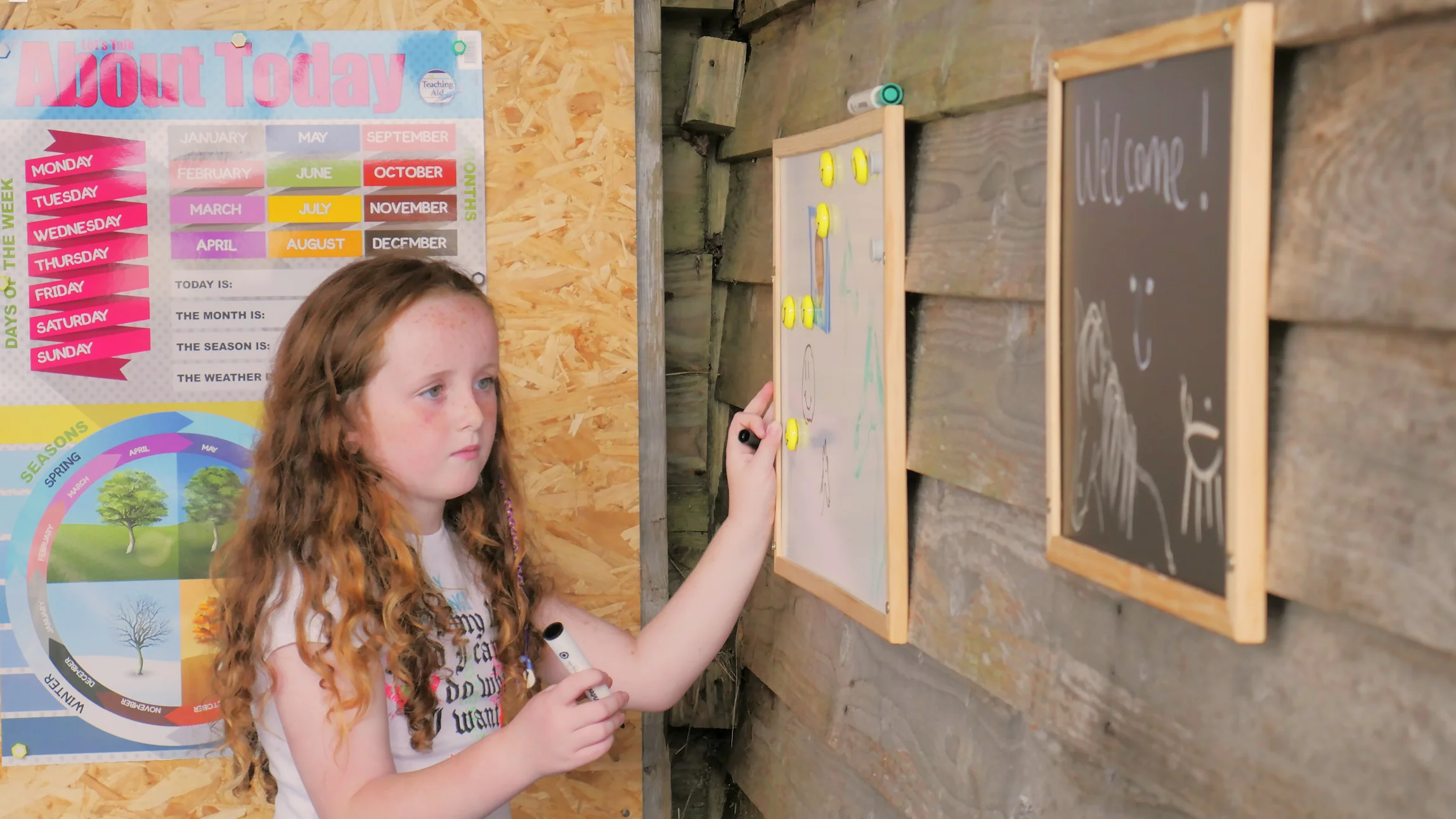 Young girl with long curly hair holding markers and drawing on a whiteboard mounted on a wooden wall inside a classroom.