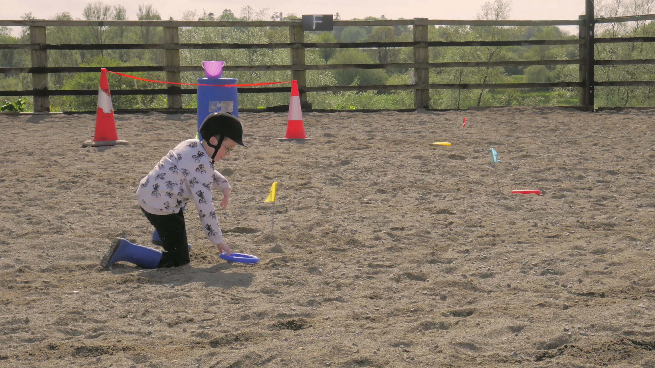 Child wearing a helmet and blue boots kneeling on sand, playing with colorful rings near small flags and traffic cones.