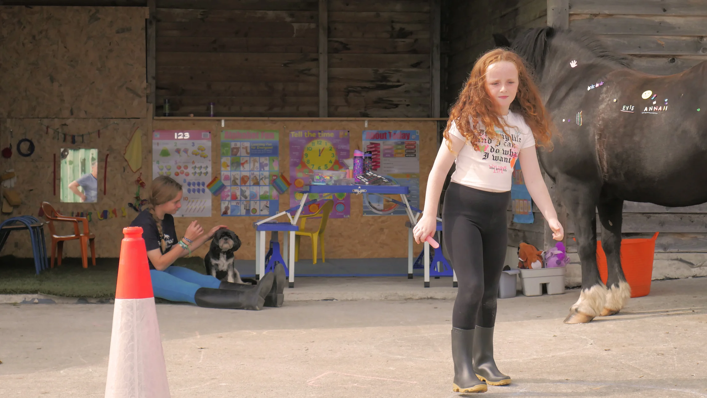 Girl with red curly hair in black leggings and boots standing near a black horse decorated with stickers, while another girl sits on the ground petting a small black and white dog in a wooden shelter with colorful educational posters.
