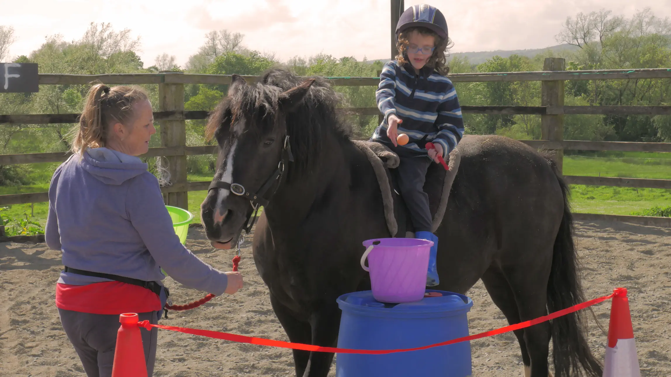 Child wearing a helmet sitting on a black horse while holding a ping pong paddle, accompanied by an adult woman holding the horse's lead rope in an outdoor fenced area.