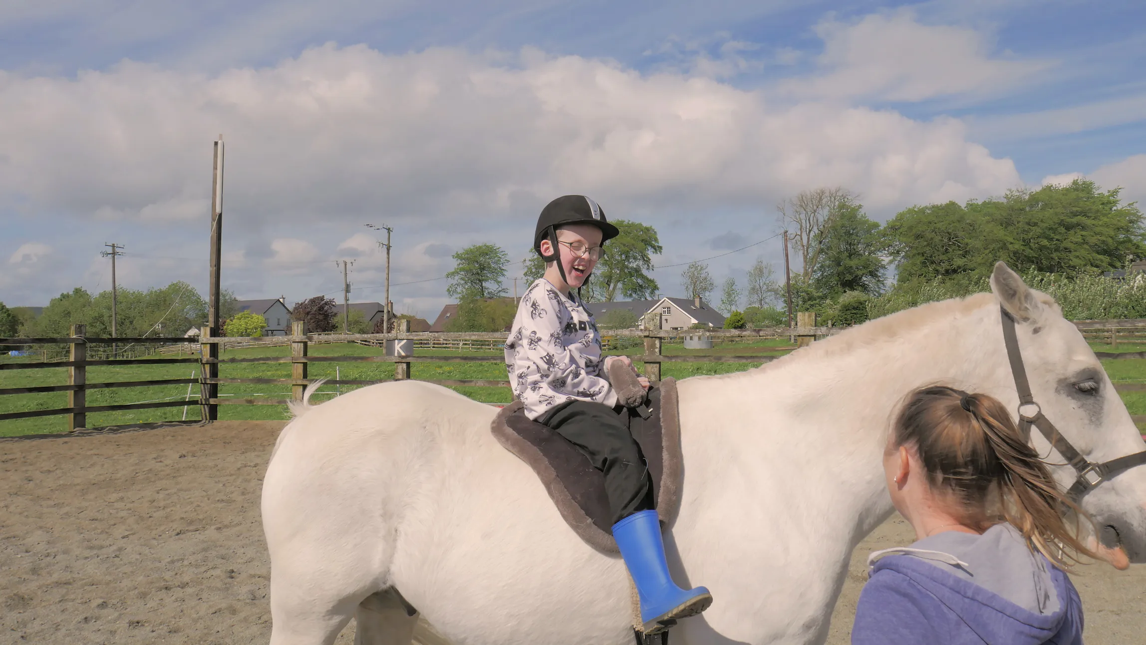 Smiling child wearing a helmet and blue boots riding a white horse with a woman holding the horse's reins.
