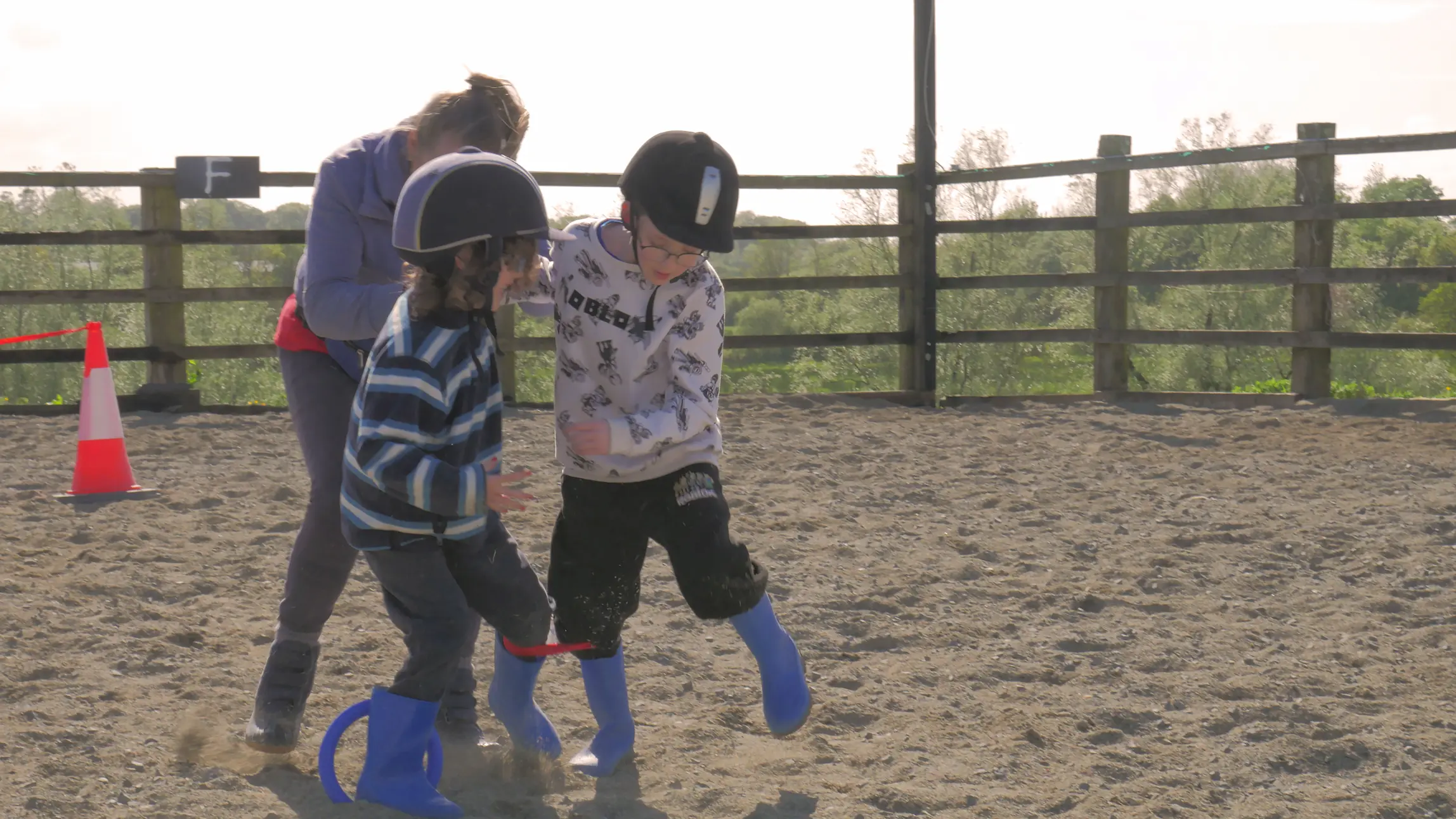 Two children wearing helmets and blue rain boots playing a three-legged race on sandy ground with an adult supervising.