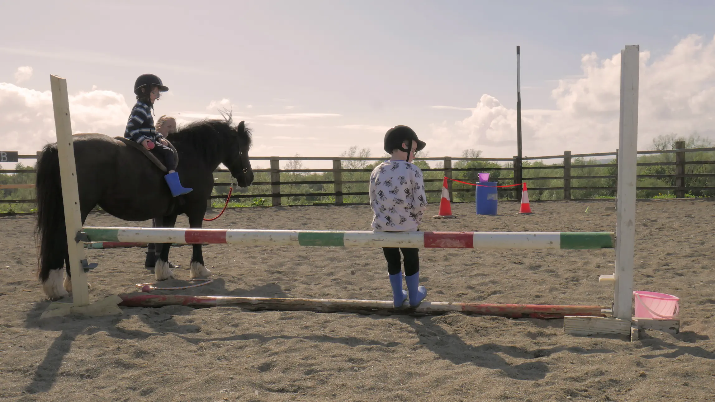 Two children wearing helmets and blue boots in a sandy horse riding arena, one sitting on a black pony and the other standing on a wooden jump pole.