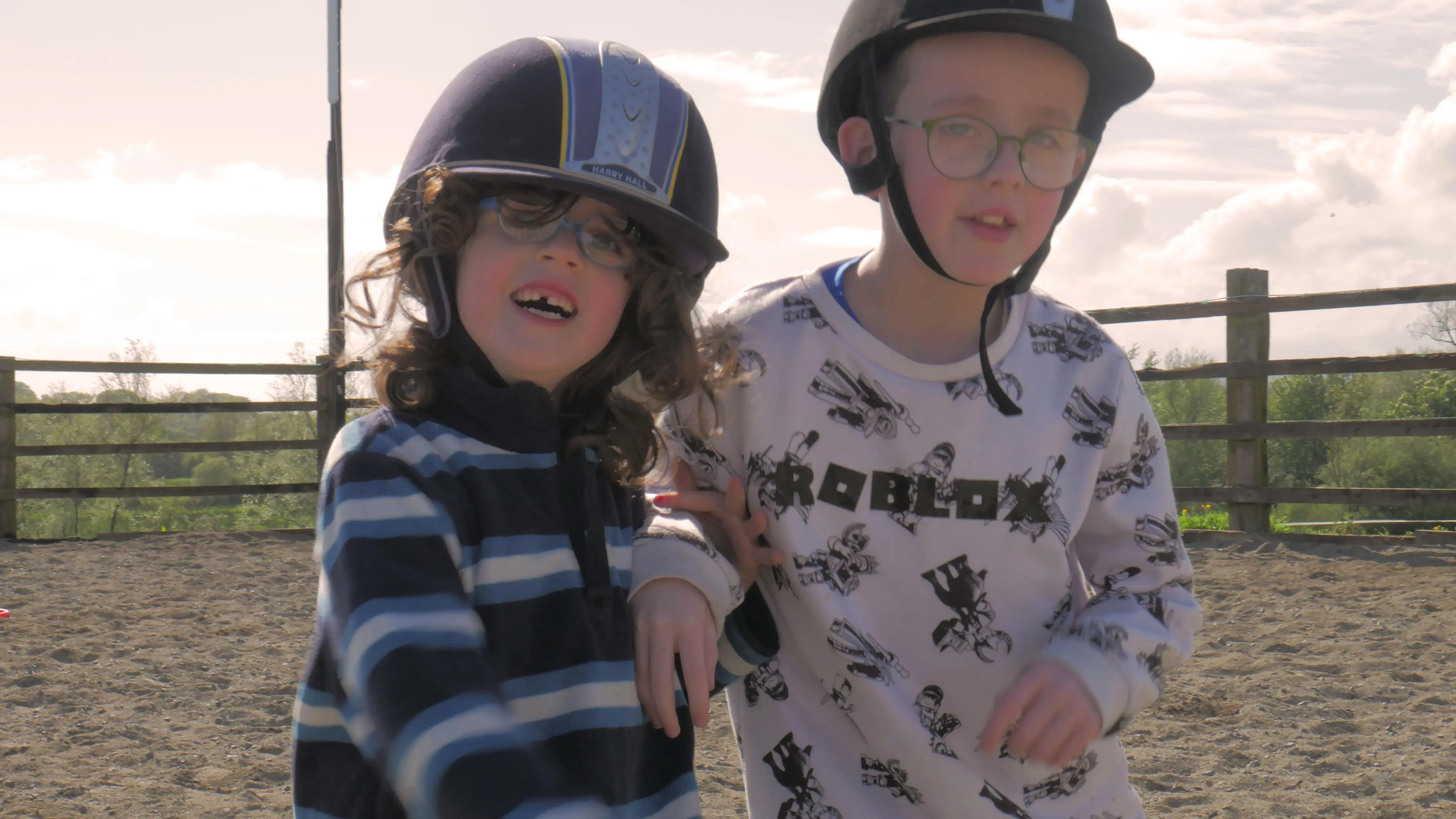 Two children wearing helmets and glasses, standing close together in an outdoor sandy area with a wooden fence and trees in the background.