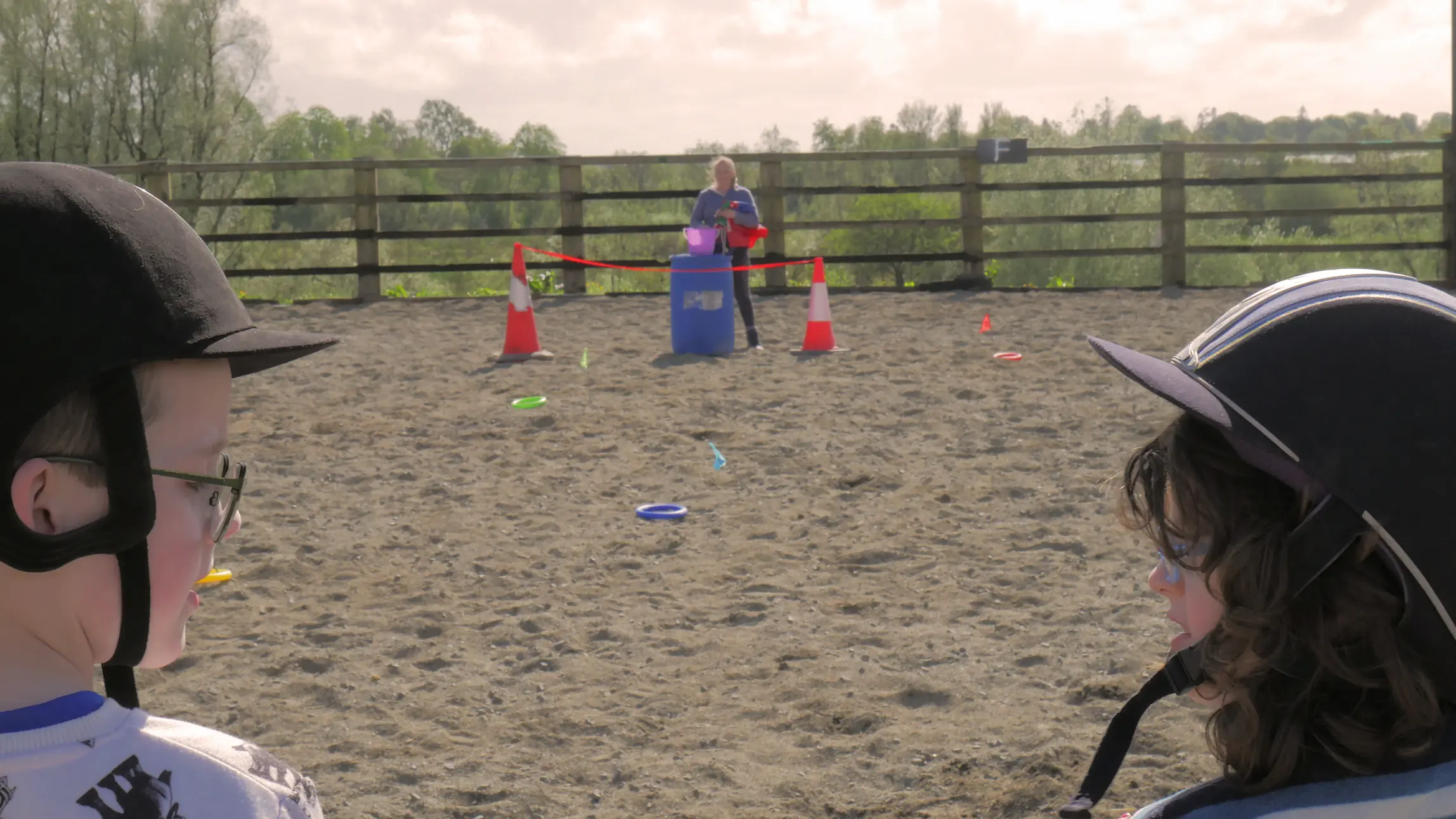 Two children wearing equestrian helmets facing a woman standing behind a blue barrel with traffic cones on a sandy ground.
