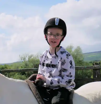 Boy wearing glasses and a black riding helmet sitting on a white horse outdoors under a partly cloudy sky.