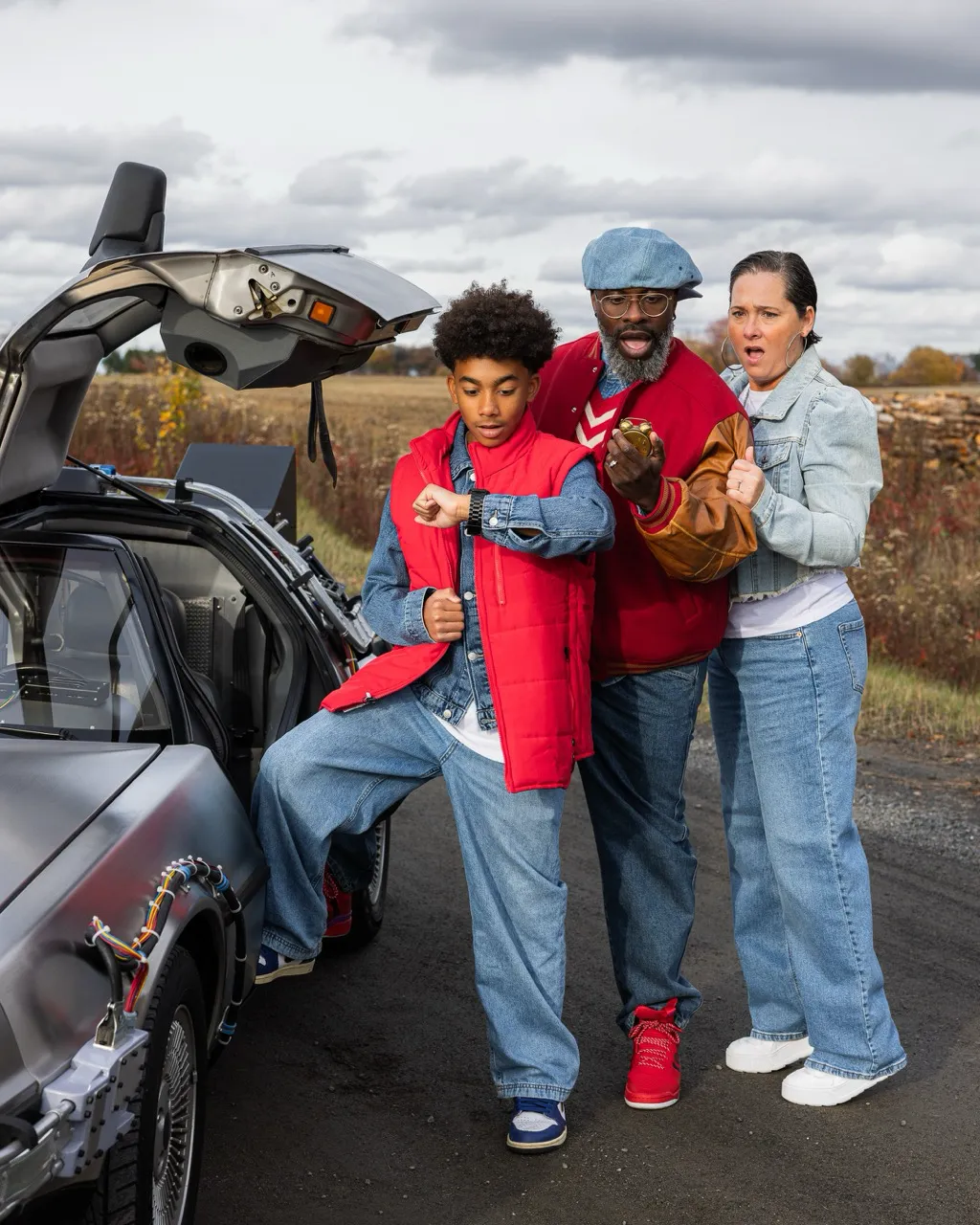 A young man in a red vest checking his smartwatch next to a man holding a stopwatch and a woman, all reacting with surprise beside a silver car with gull-wing doors open.