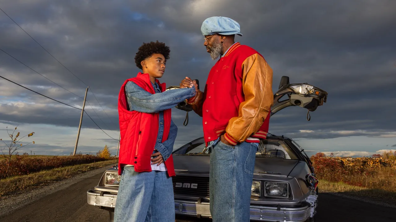 Two men dressed in red jackets shake hands in front of a DeLorean car with gull-wing doors open on a cloudy day.