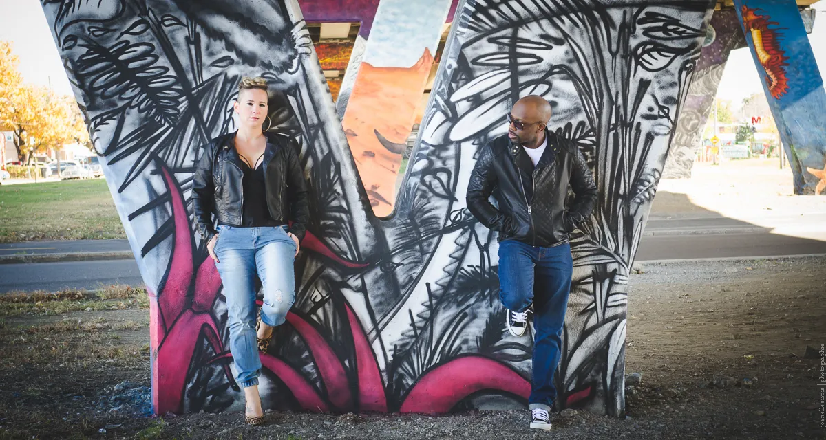 Man and woman in leather jackets leaning against graffiti-covered concrete pillars under a bridge.
