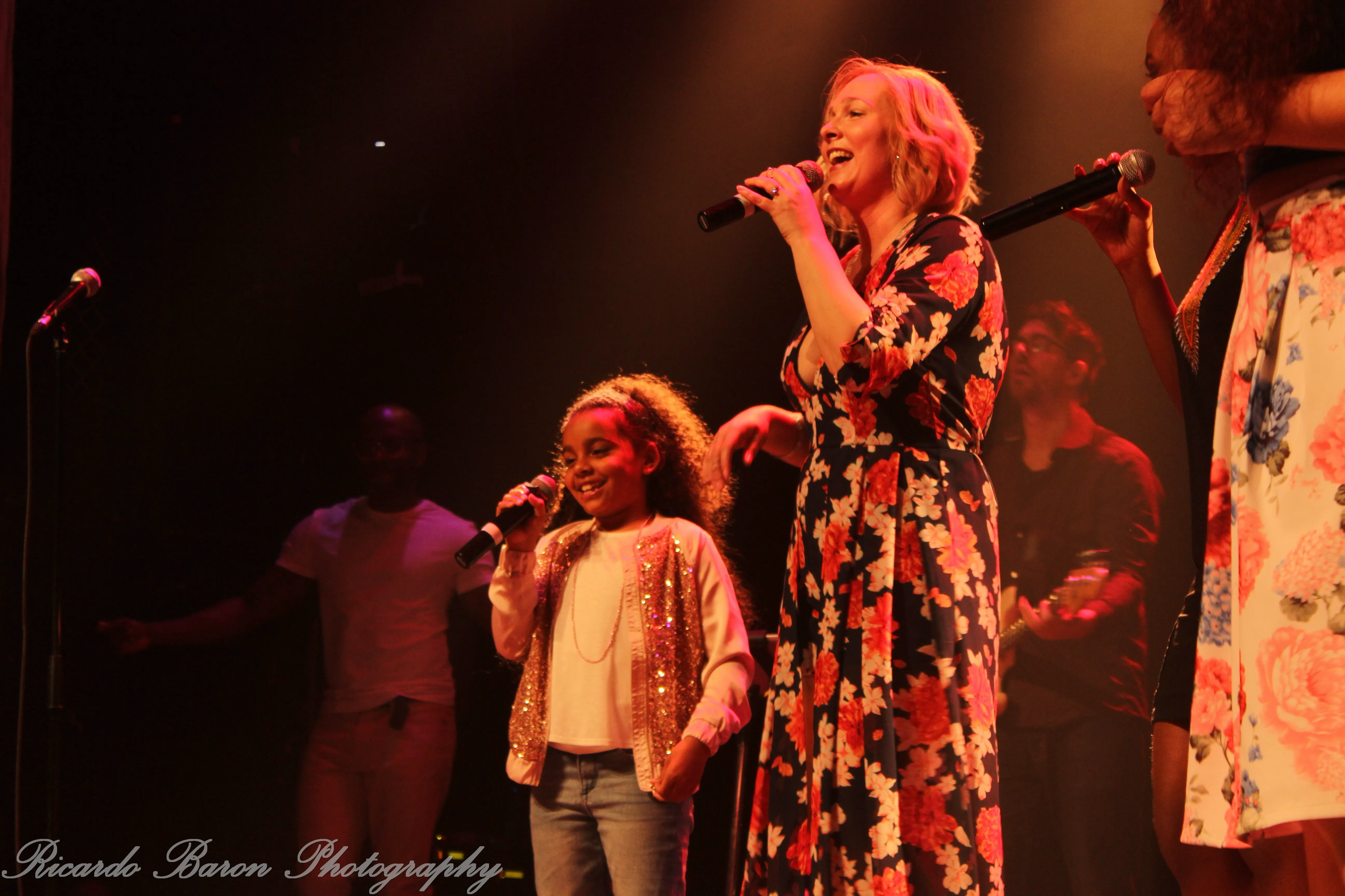 A young girl and a woman in a floral dress singing into microphones on stage with musicians in the background.