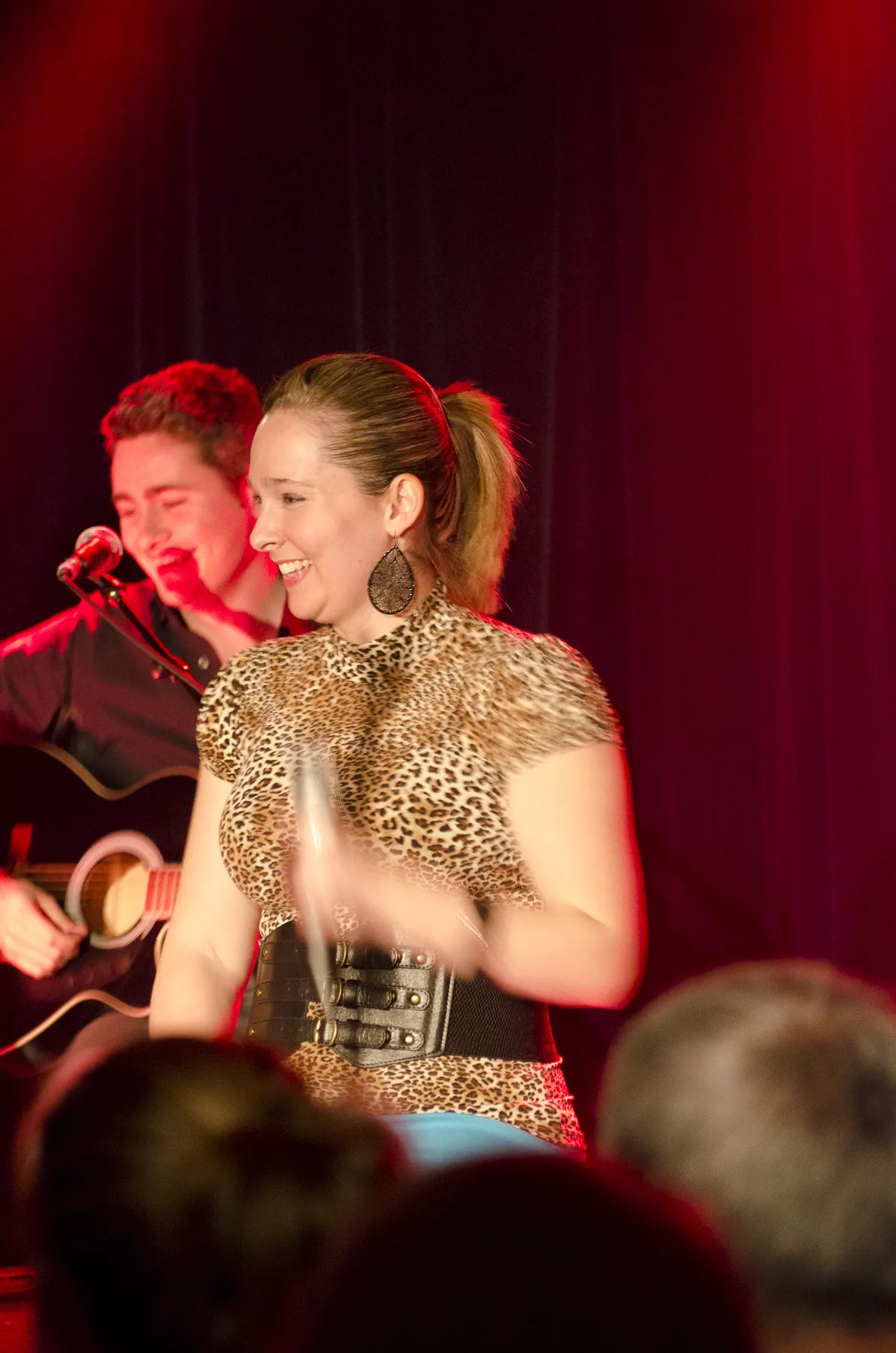 Smiling female singer in a leopard print top holding a microphone, with a guitarist in the background on stage.