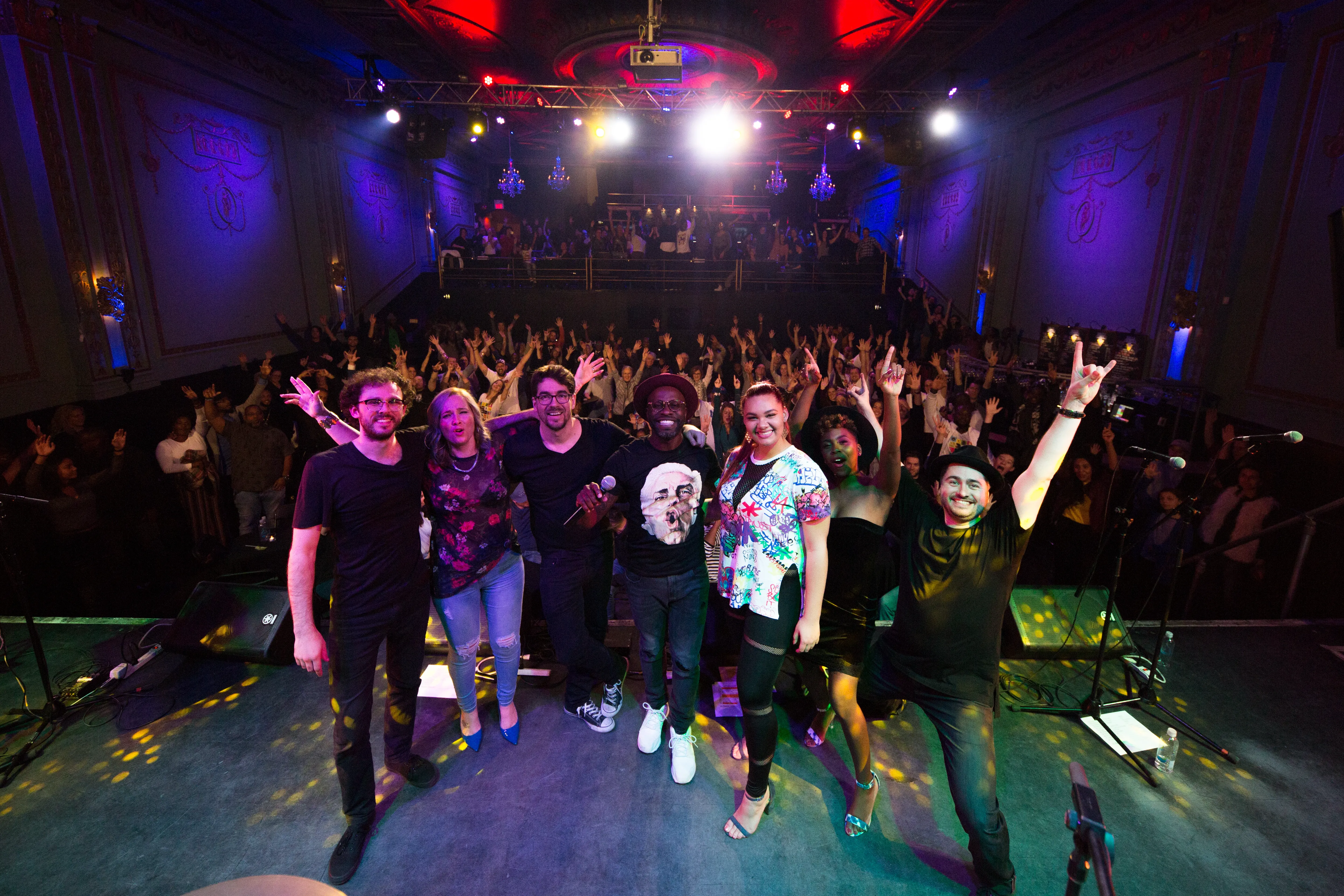 A diverse group of seven performers posing on stage in front of a lively audience in a dimly lit theater with colorful stage lights.