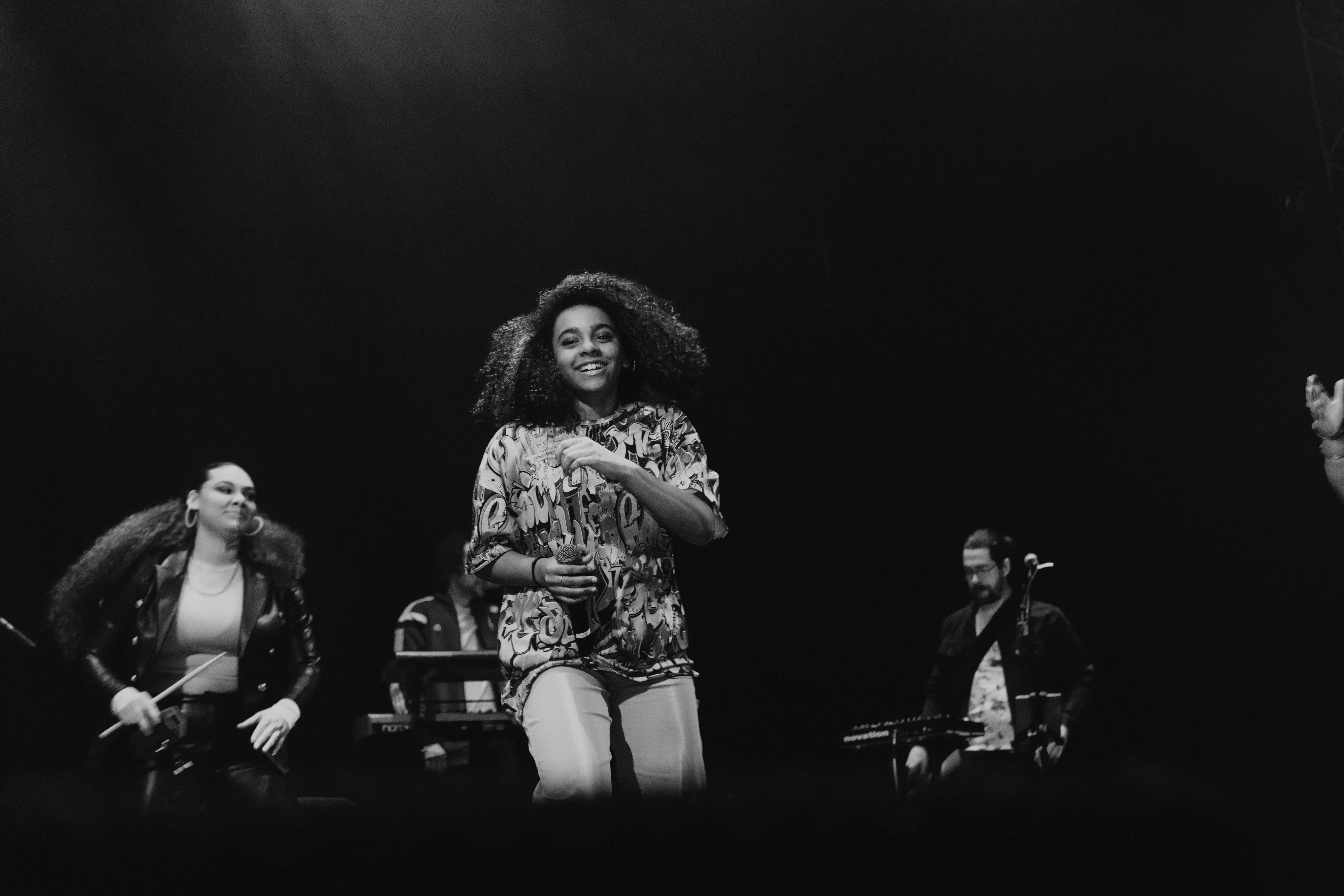 Black and white photo of a female singer smiling and holding a microphone on stage with musicians in the background.