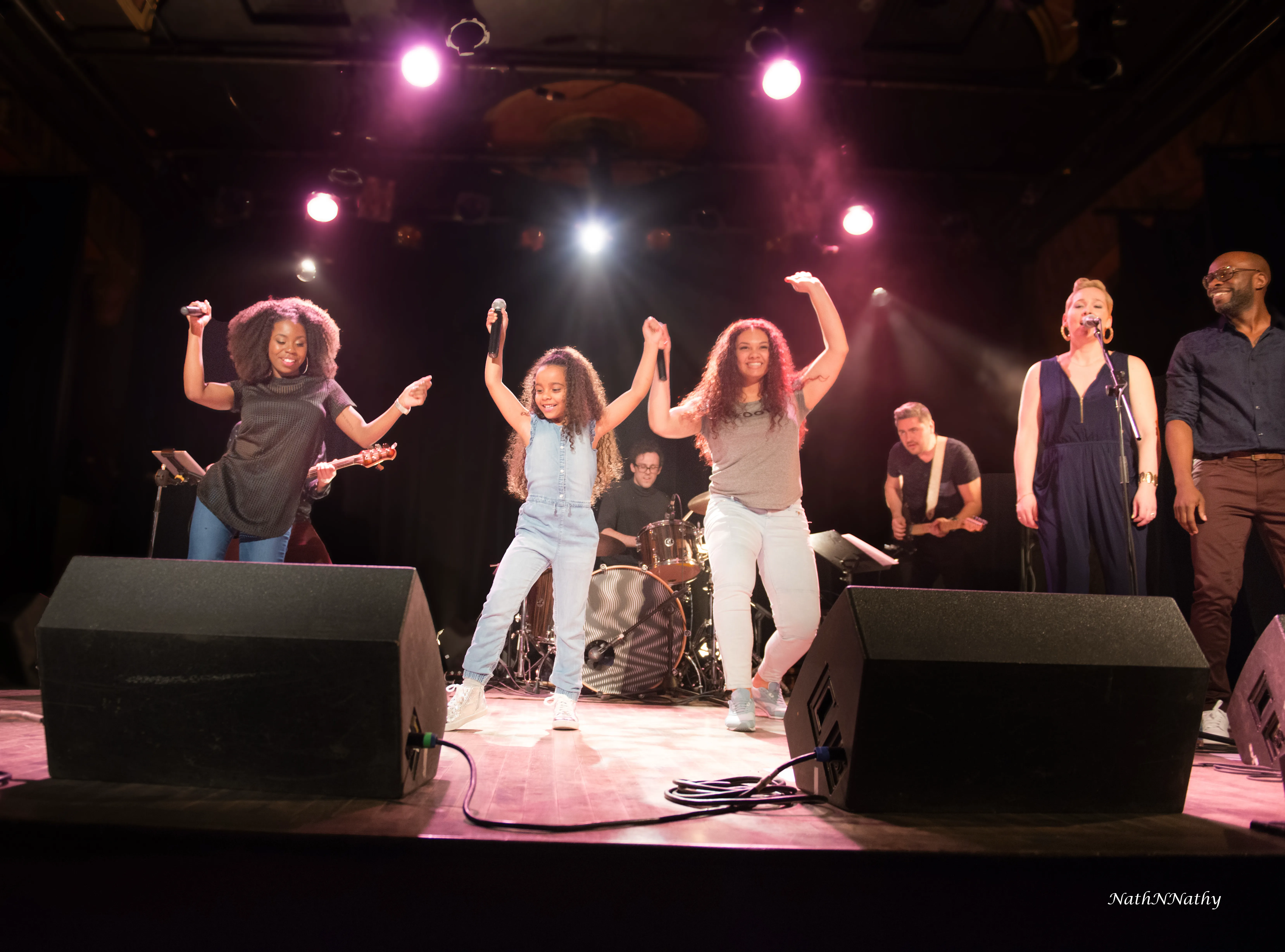 A diverse group of seven musicians and singers performing on stage with bright stage lights, including a drummer in the back and vocalists dancing with microphones in the front.